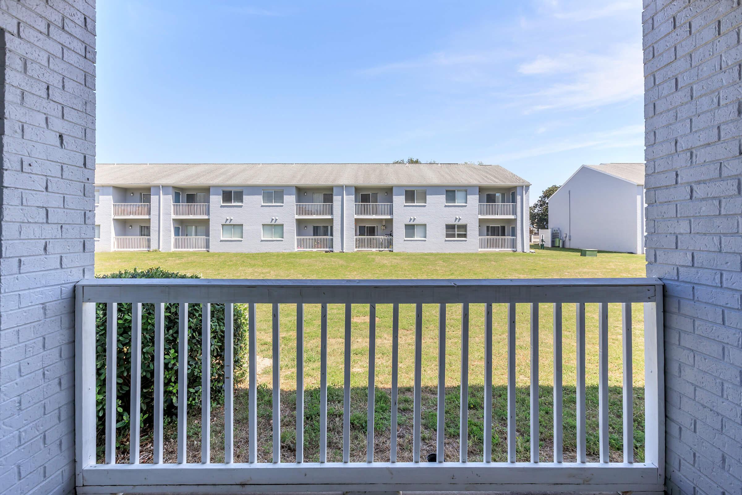 View from a balcony overlooking a grassy area with two multi-story apartment buildings in the background. The sky is clear and blue, providing a bright and open atmosphere. The railing of the balcony is visible in the foreground, adding depth to the scene.