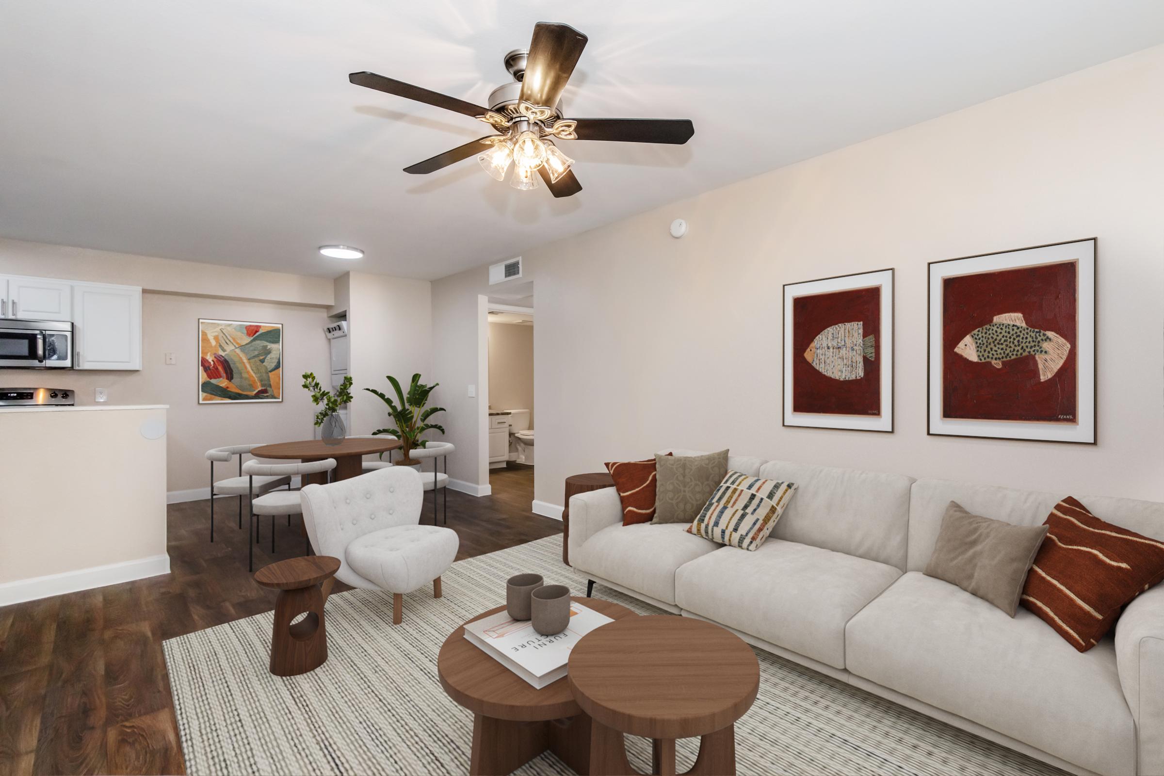 A modern living room featuring a light-colored sofa with decorative pillows, a round coffee table, and an area rug. A ceiling fan with lights hangs above, while artwork is displayed on the walls. In the background, a dining area is visible with a table and chairs, and a potted plant adds a touch of greenery.