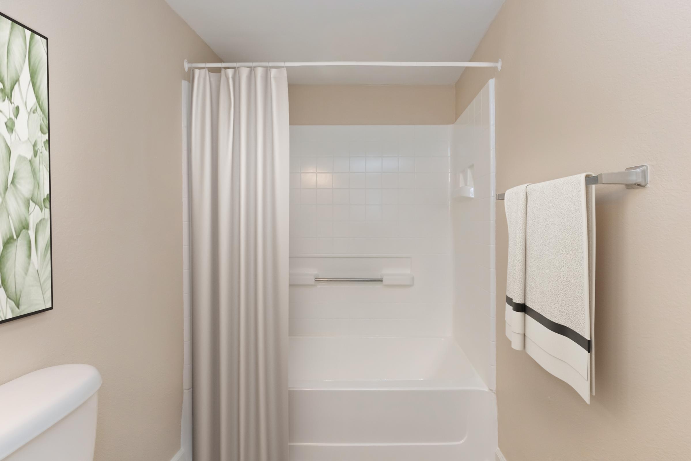 A clean and modern bathroom featuring a white bathtub with a shower curtain, beige walls, and a neatly hung towel on a rack. An artwork with green leaves is visible on the wall. The overall design is simple and inviting.