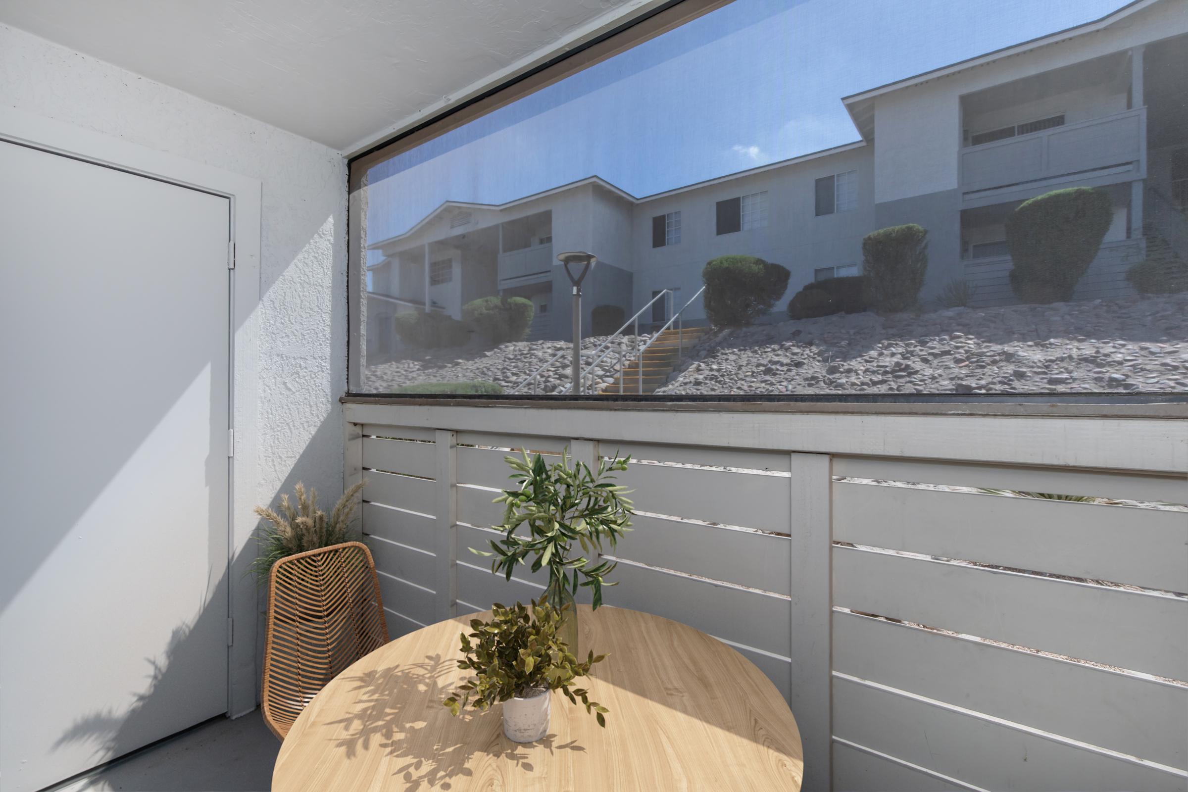 A bright balcony scene featuring a small round wooden table with a plant centerpiece. In the background, a multi-story apartment building can be seen through a protective screen, alongside landscaped shrubbery and an outdoor light fixture. Natural light illuminates the space.