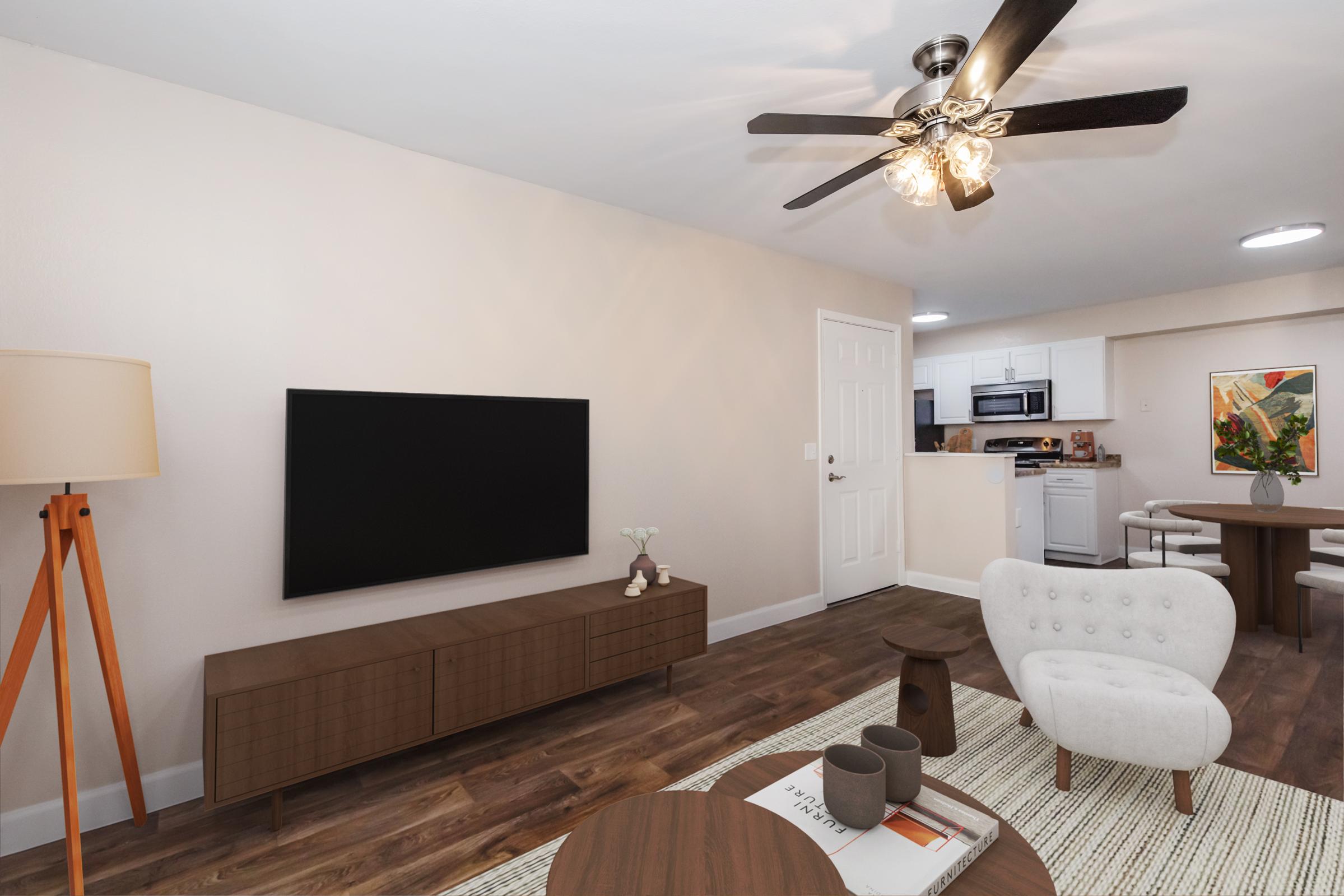 A modern living room featuring a flat-screen TV mounted on a light-colored wall, a stylish wooden media console, a white accent chair, a round coffee table, and an area rug. A ceiling fan with lights hangs above, and in the background, there's a dining area connected to a kitchen with white cabinets.