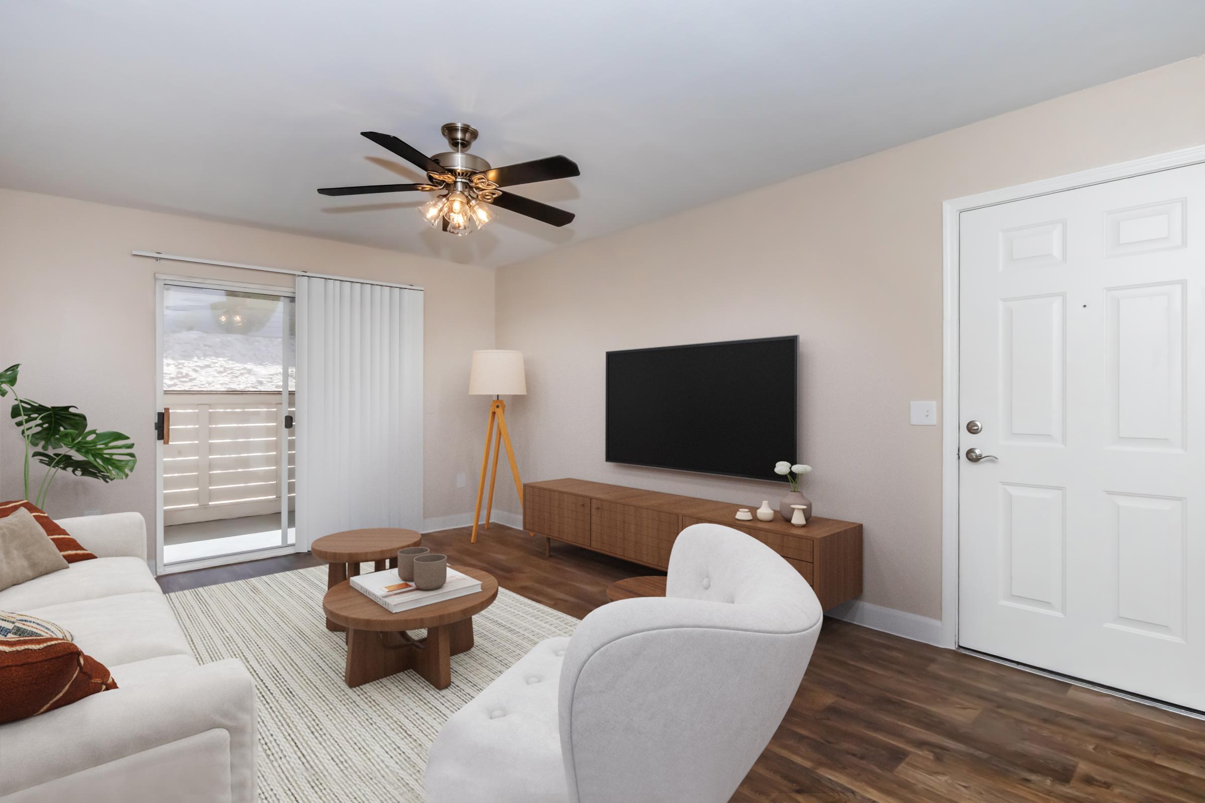 A cozy living room featuring a light-colored sofa with decorative pillows, a wooden coffee table, and a TV mounted on the wall. A ceiling fan provides added comfort, while a large window with sliding glass doors allows natural light in. A floor lamp and indoor plant add warmth to the space.