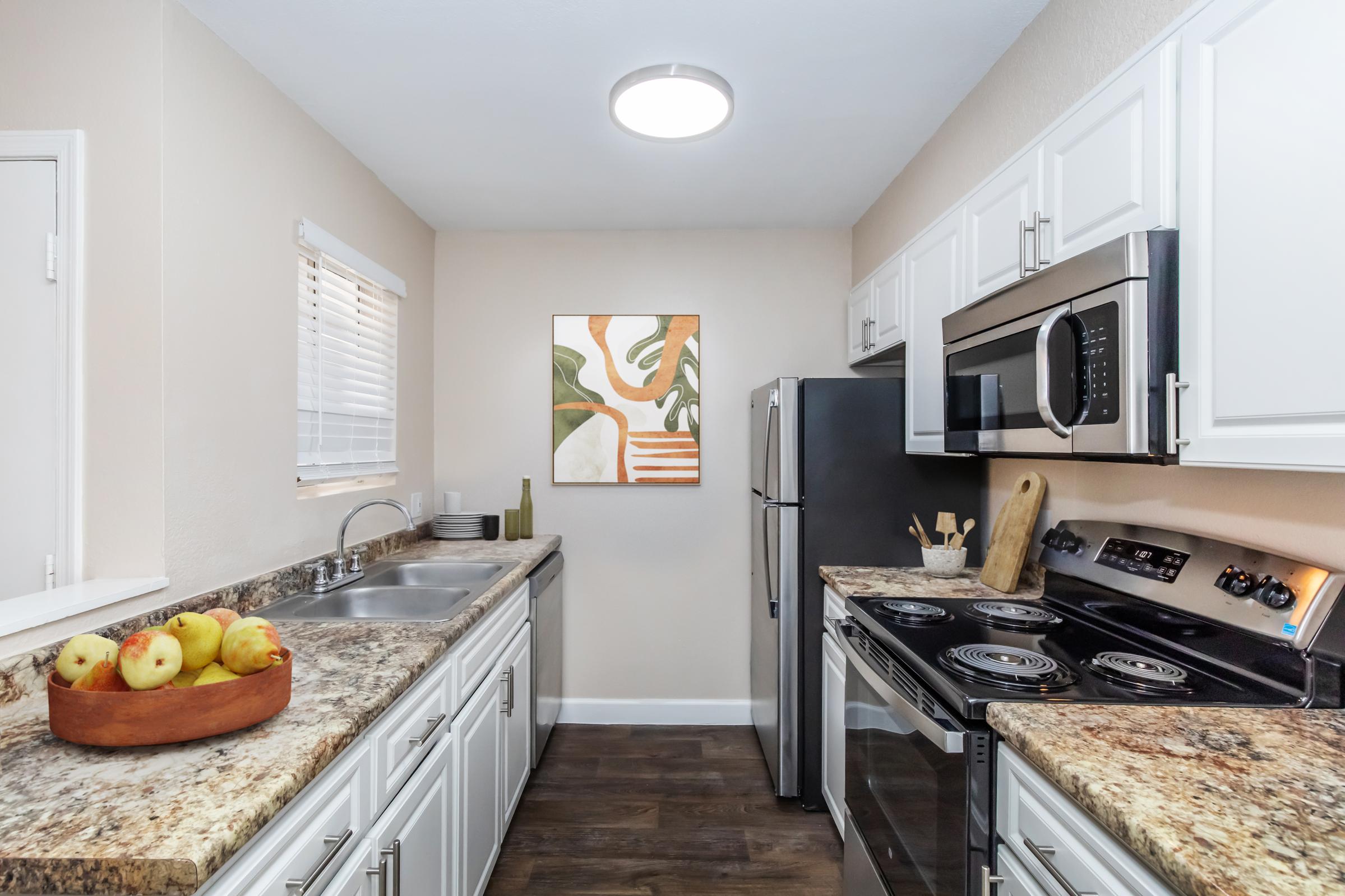 A modern kitchen featuring white cabinets, a stainless steel refrigerator, and an oven. The countertop is covered with granite, and a bowl of apples sits on the counter. A piece of wall art is displayed, and there are kitchen utensils and appliances visible. Natural light is streaming in through a window.