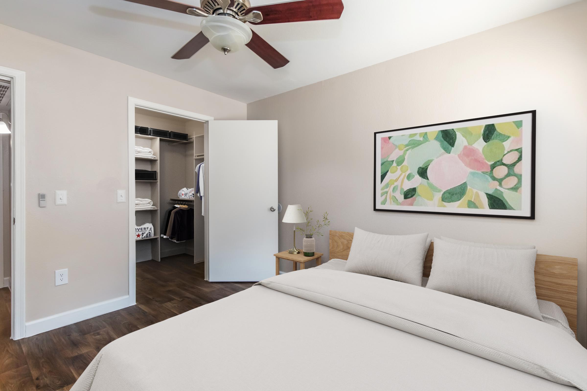 A cozy bedroom featuring a queen-sized bed with light-colored bedding, a small bedside lamp, and a framed abstract artwork on the wall. There's a closet with open doors, showcasing organized shelves and clothing. The walls are painted a soft beige, and a ceiling fan provides ventilation.