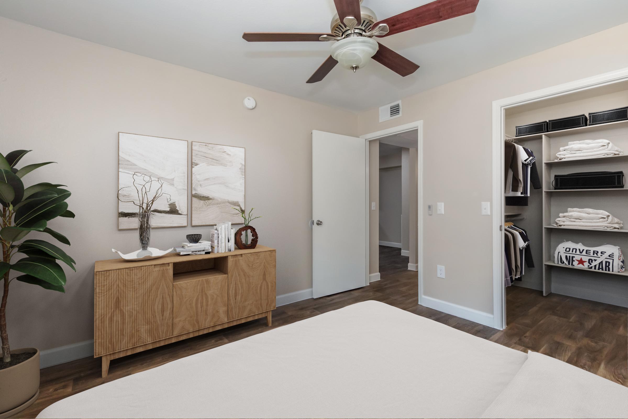 A well-decorated bedroom featuring a light-colored wall, a ceiling fan, and a wooden dresser with decorative items. To the right, there is an open closet showcasing stacked towels and clothing. A potted plant adds a touch of greenery, while two abstract paintings hang above the dresser, enhancing the room's aesthetic.