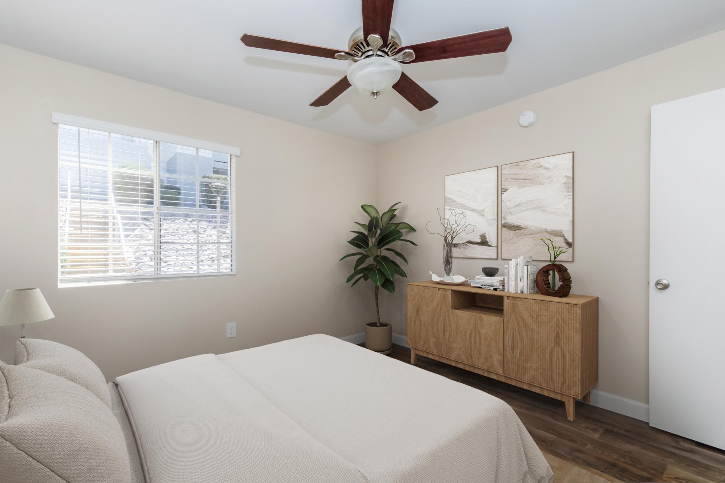 A cozy bedroom featuring a neutral color palette. There is a queen-sized bed with a beige comforter, a ceiling fan with wooden blades, a window letting in natural light, a decorative plant in the corner, and stylish wall art above a wooden cabinet with decorative items.