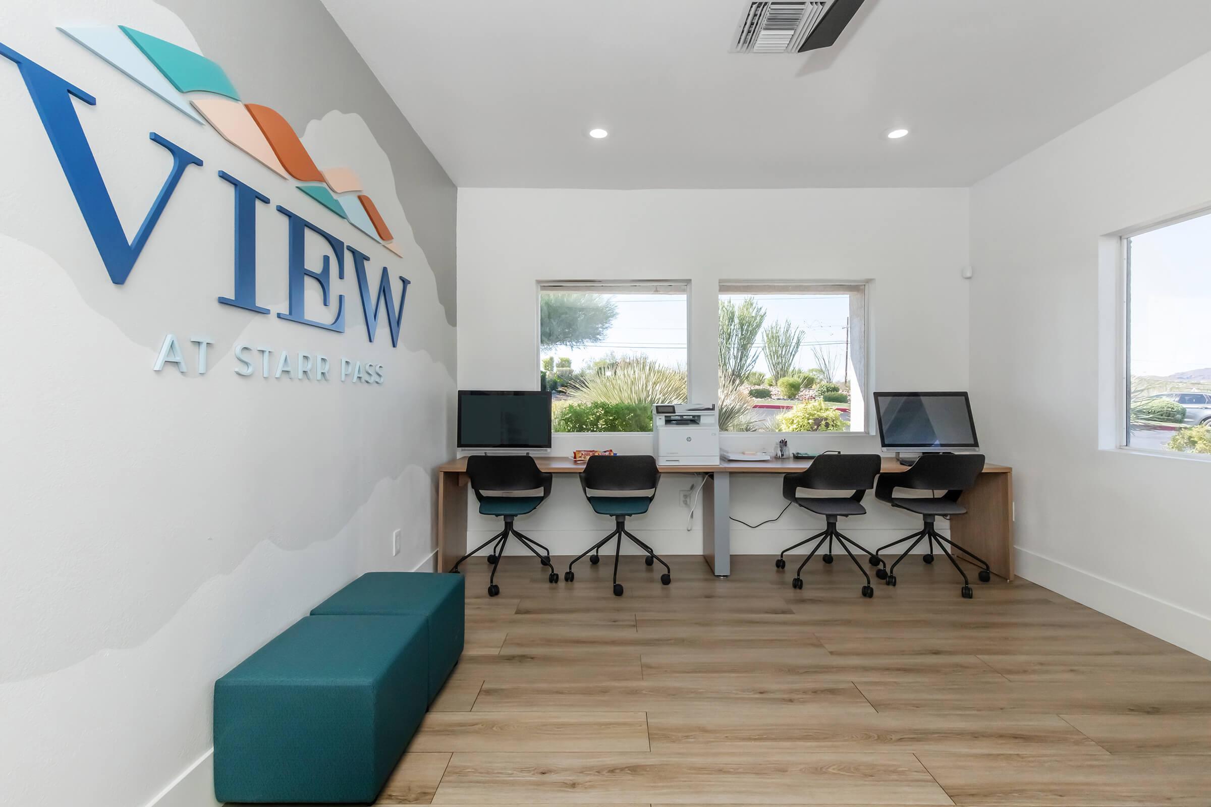 A modern office space featuring a sign that reads "VIEW" on the wall. There are three workstations with desktop computers and chairs, and a teal bench is positioned against the wall. Large windows provide natural light and a view of greenery outside. The flooring is light-colored wood.