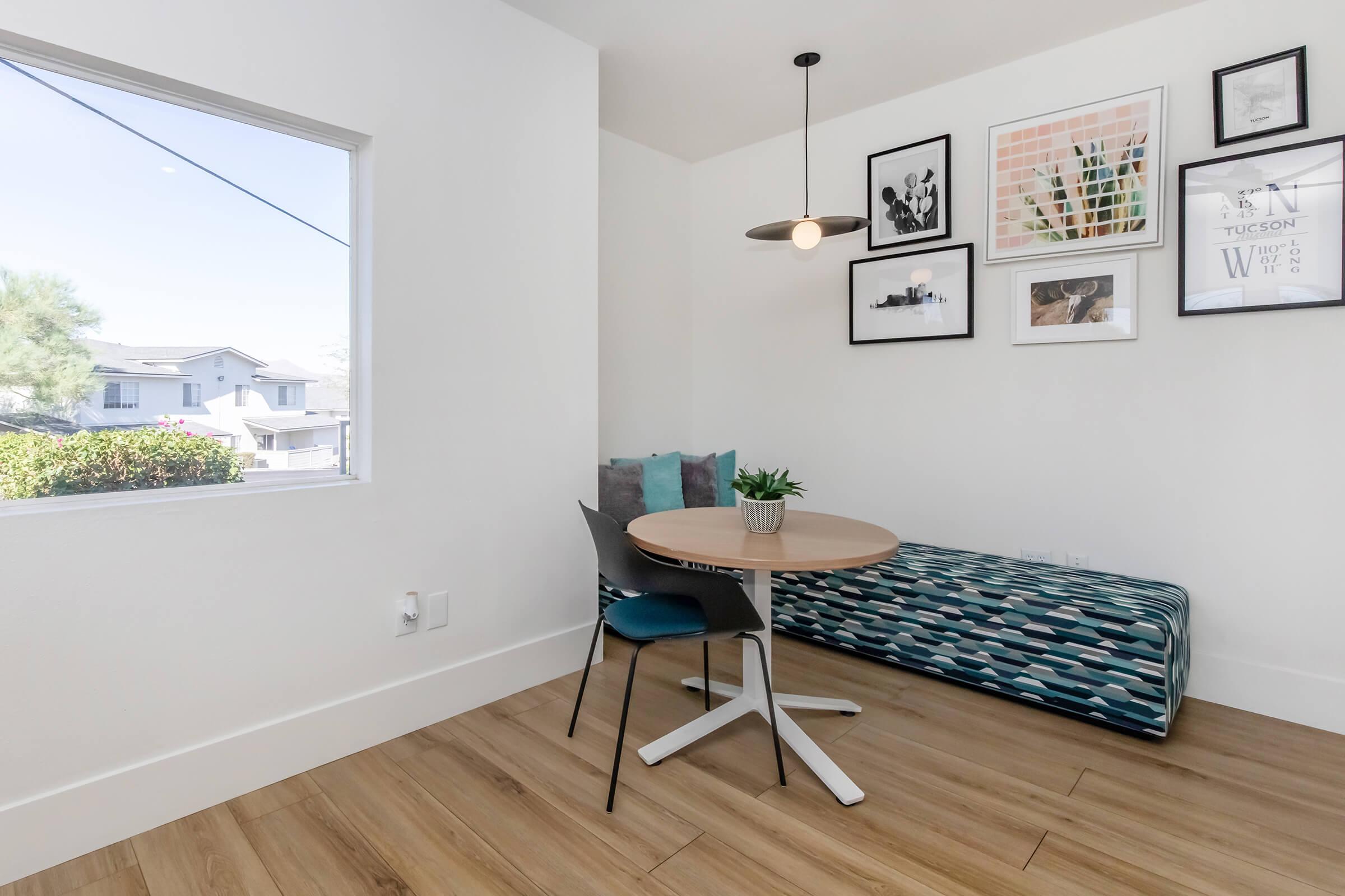 A cozy dining nook featuring a circular wooden table with a modern base, two chairs, and a patterned bench against the wall. The wall is decorated with various framed photos and artwork, and a small potted plant sits on the table. A window lets in natural light, enhancing the inviting atmosphere.