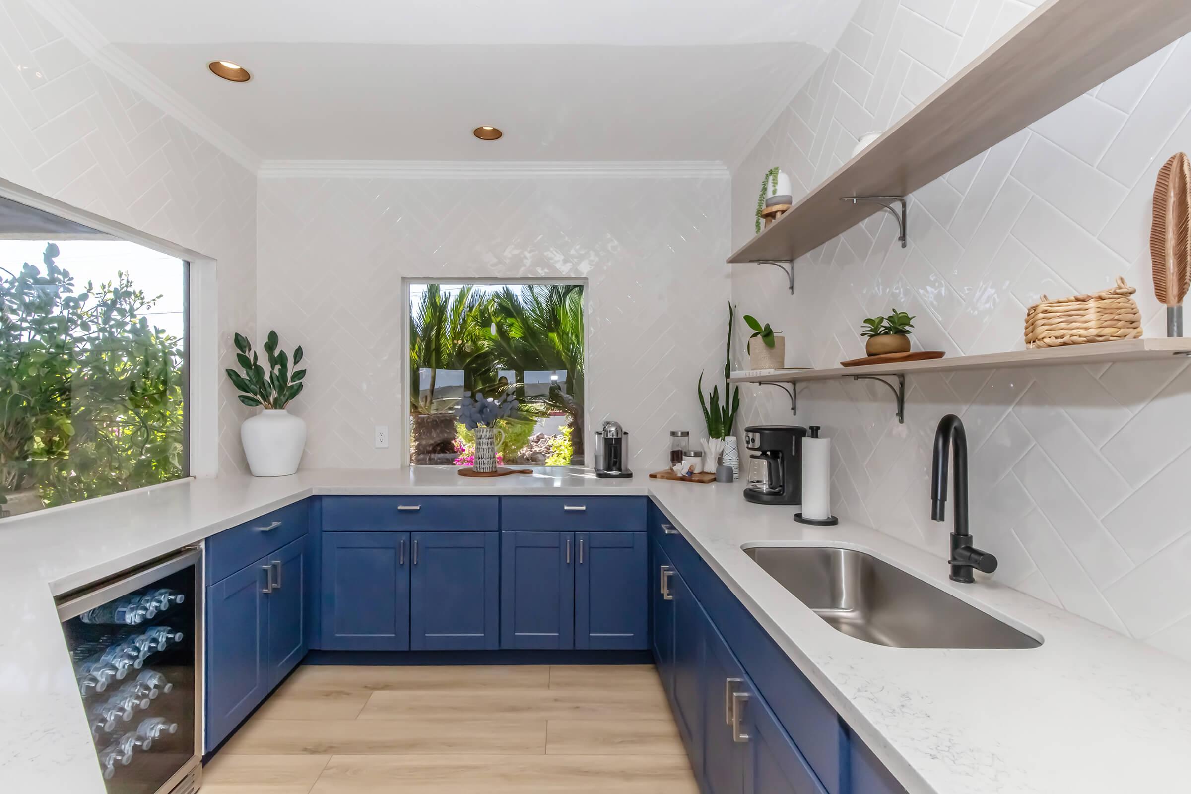 Modern kitchen featuring navy blue cabinetry, white countertops, and open shelving. Plants adorn the shelves, and a window offers a view of greenery outside. A coffee maker and wine cooler are visible, contributing to a stylish and functional space. The flooring is light wood, enhancing the bright atmosphere.