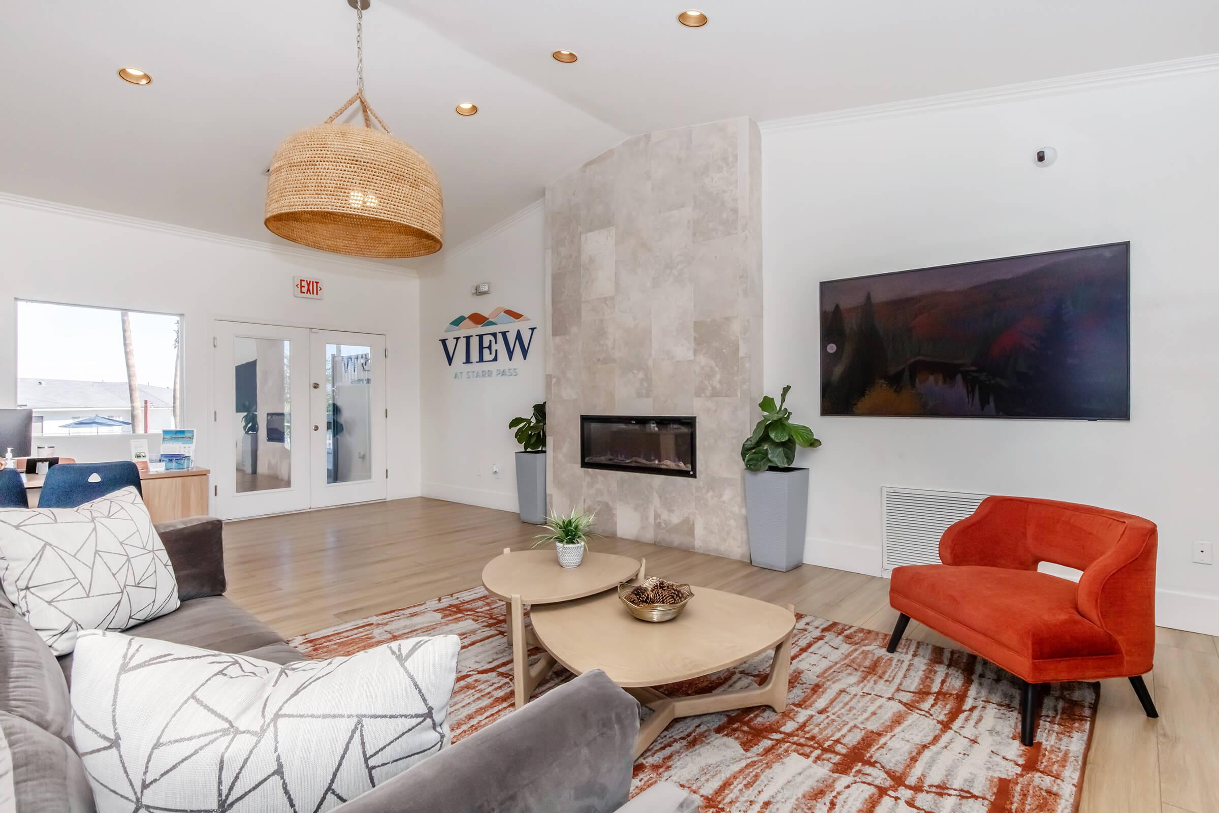 Modern living room with a cozy atmosphere, featuring a gray sectional sofa adorned with patterned cushions, a round coffee table, and an orange armchair. The space includes a stylish stone fireplace, a large wall-mounted TV, and potted plants, with a welcoming sign that says "VIEW" in the background.