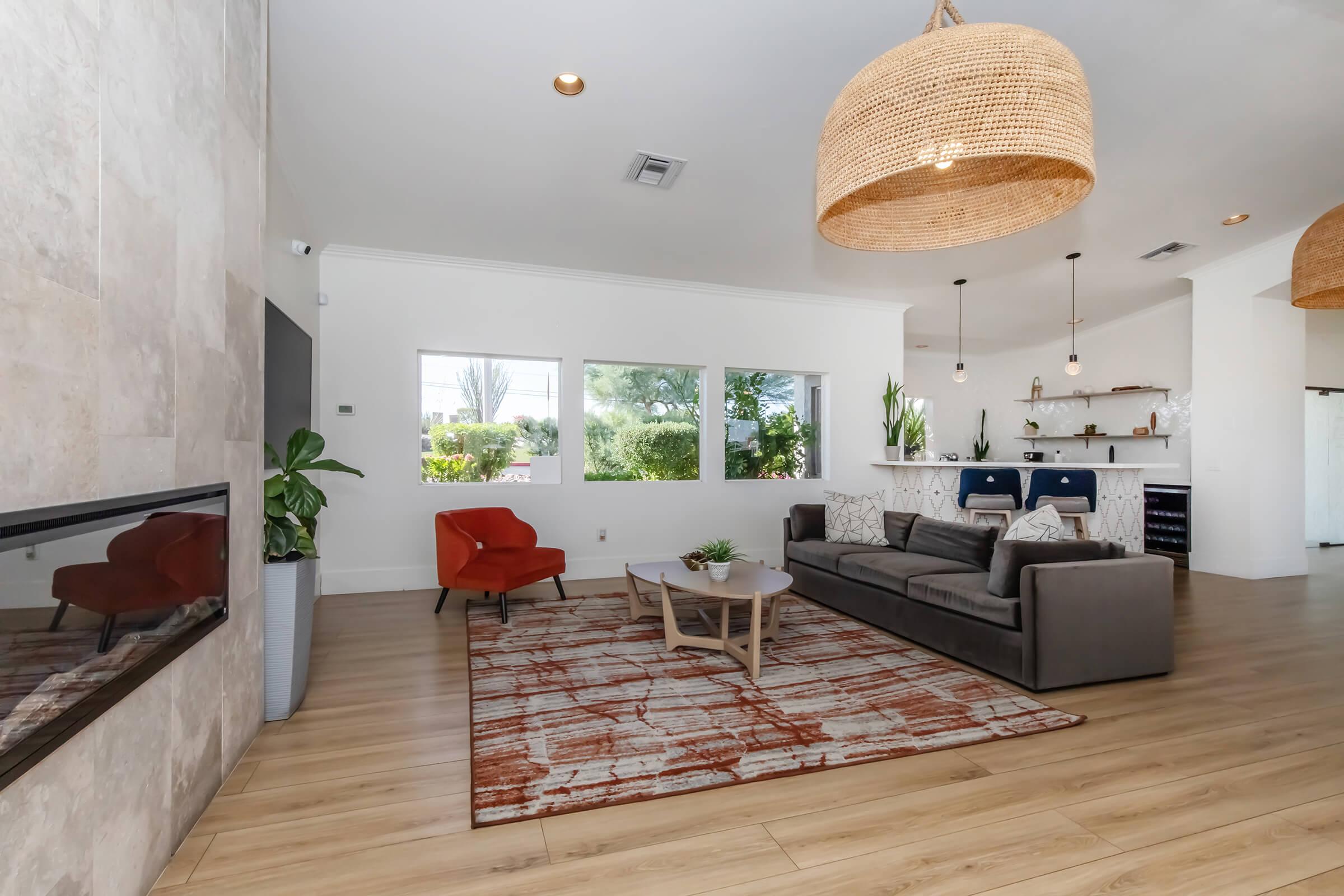 A modern living room featuring a gray sofa with cushions, an orange accent chair, and a wooden coffee table on a patterned area rug. Large windows provide natural light, and there are plants in the space. The interior has a neutral color palette with stylish hanging light fixtures.