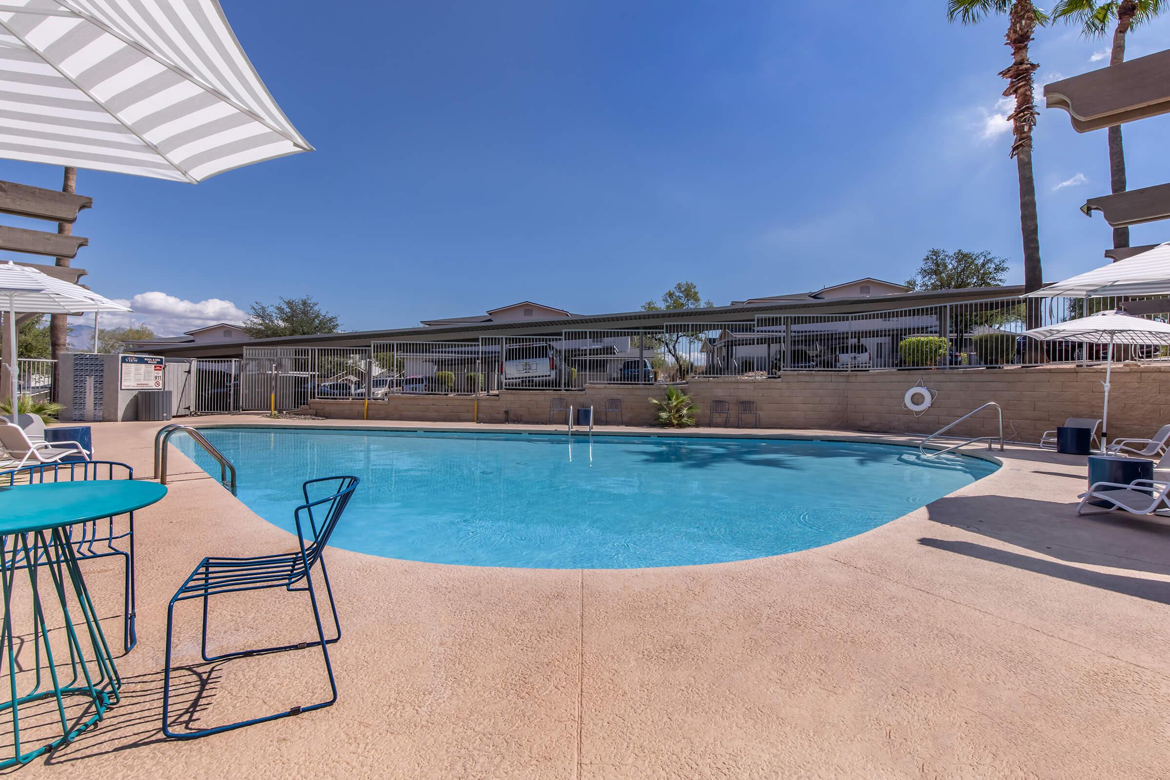 A clear blue swimming pool surrounded by sun loungers and tables with umbrellas. Palm trees line the area, and a residential building can be seen in the background under a bright blue sky. The scene conveys a relaxing outdoor atmosphere perfect for leisure and enjoyment.