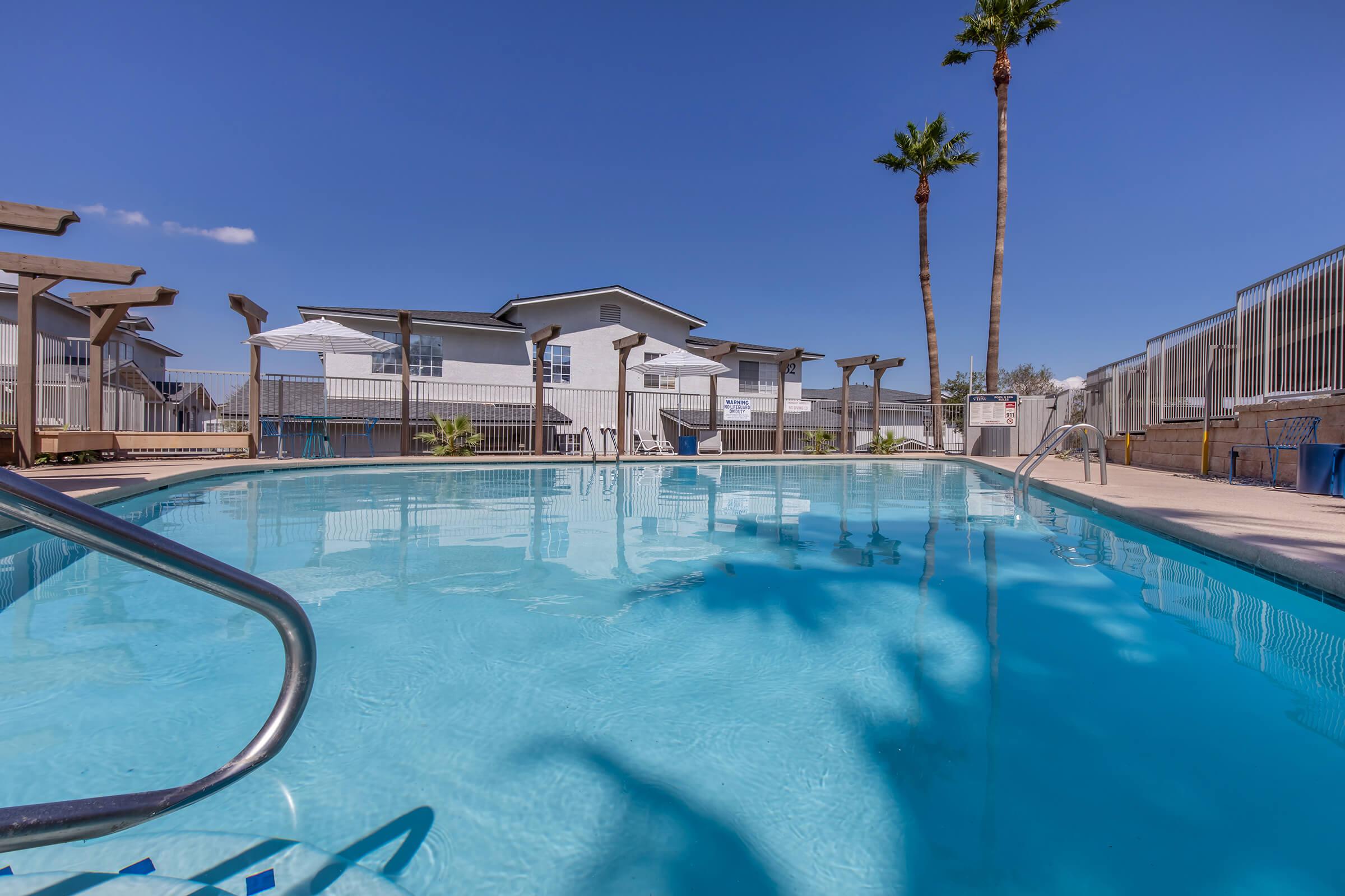 A clear swimming pool with blue water reflecting the sky, edged by a concrete area. Two tall palm trees stand in the background, alongside a modern building with multiple windows. The scene is set under a bright blue sky, creating a relaxing atmosphere.