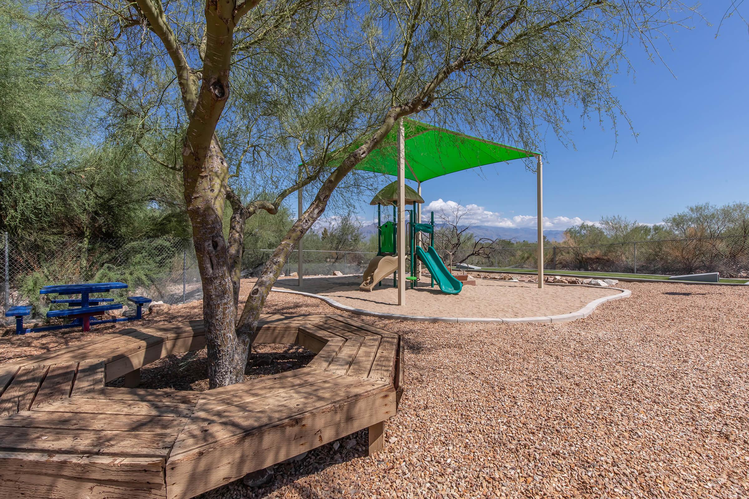 A playground area featuring a green shade structure over a slide and climbing frame. Surrounding the playground are gravel and a wooden seating area shaped like an octagon, along with a tree providing additional shade. Blue picnic tables are visible in the background against a clear blue sky.