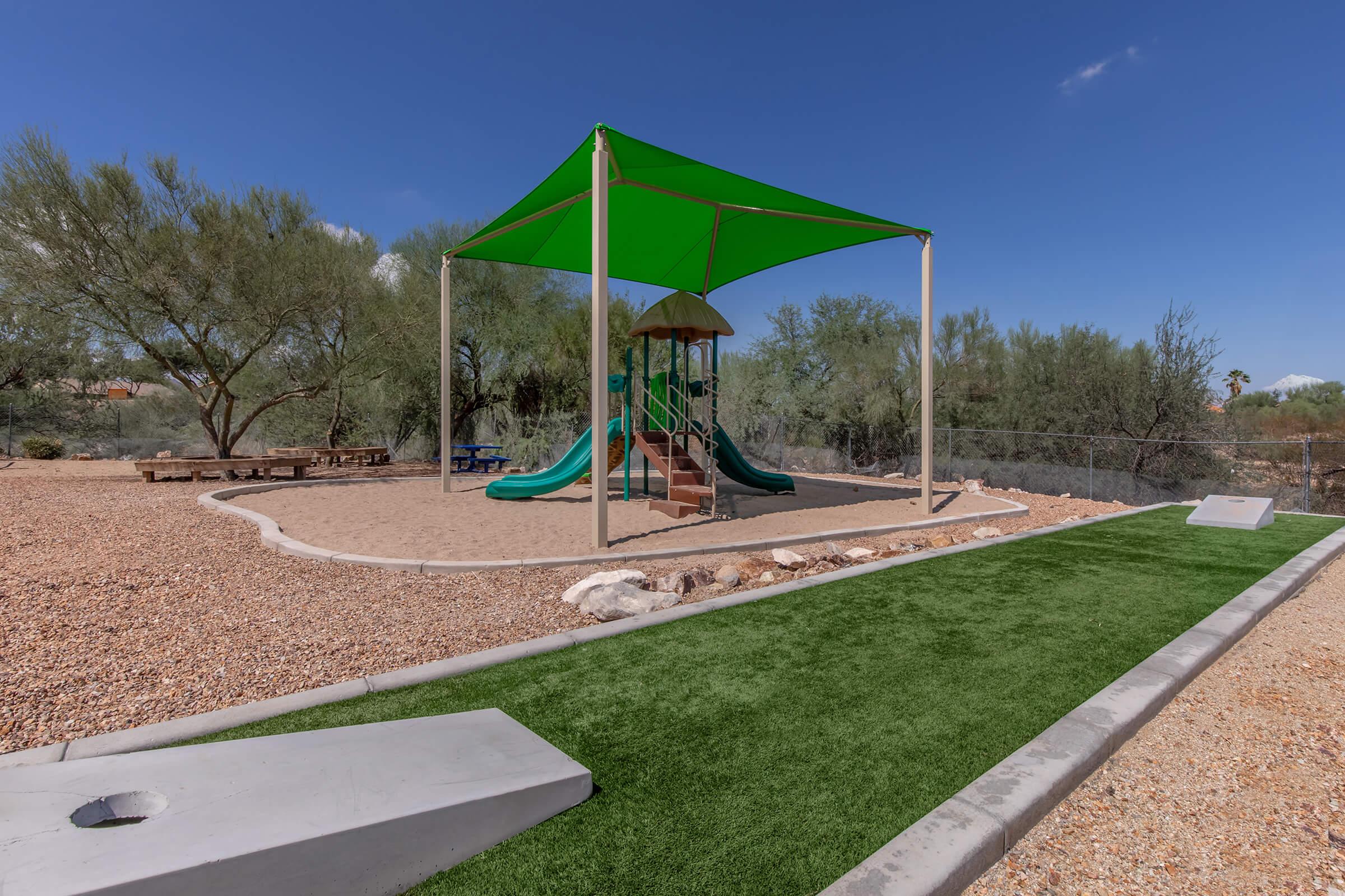 A playground with a green shade structure covering a slide and climbing equipment, surrounded by sandy ground and desert vegetation. There are benches nearby and a grassy area leading to a stone pathway. The scene is set under a clear blue sky.