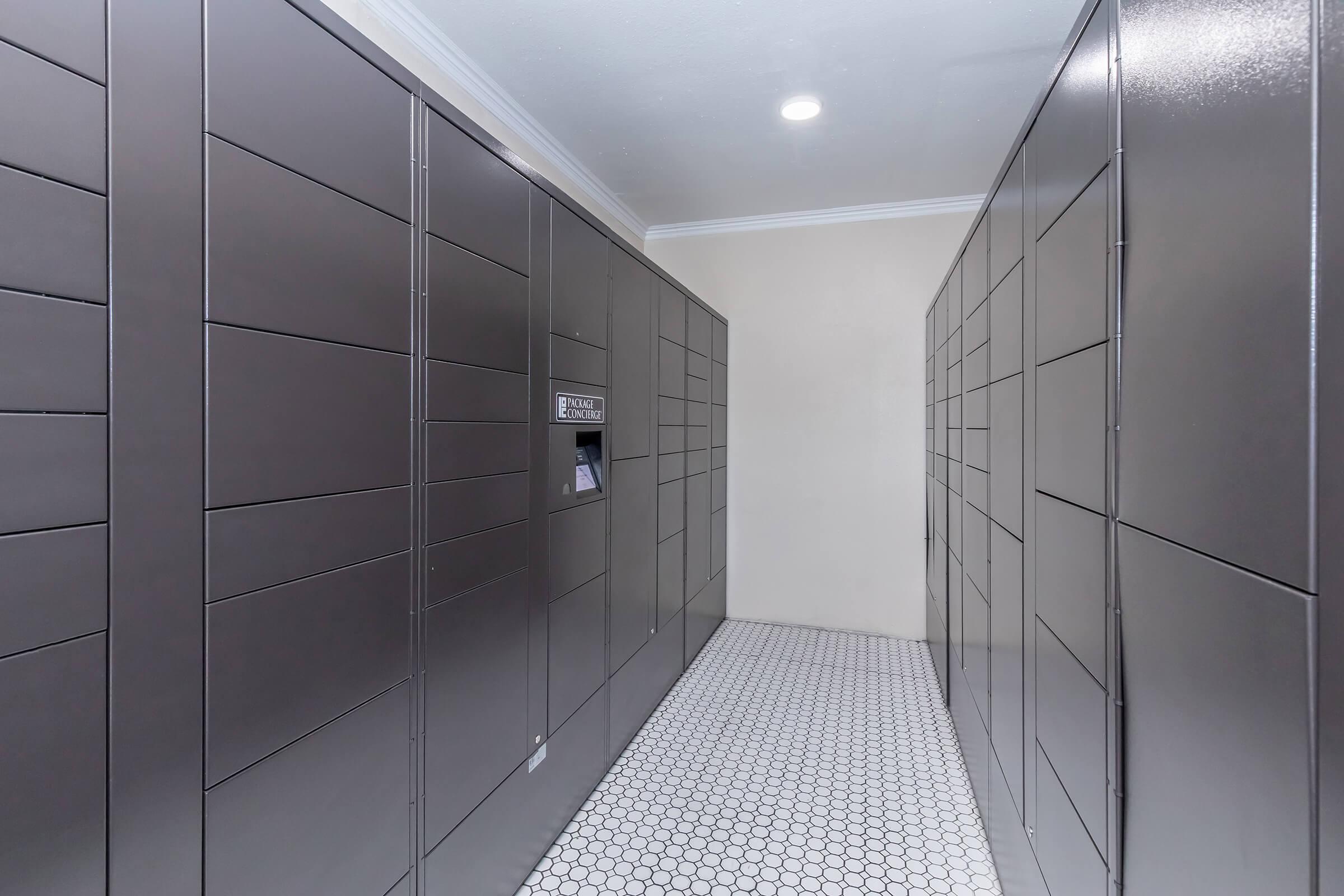 Interior view of a modern locker room with grey lockers lining the walls. The hallway is well-lit with a light fixture on the ceiling and features a patterned tile floor. A digital screen can be seen on one of the lockers, likely for accessing packages.