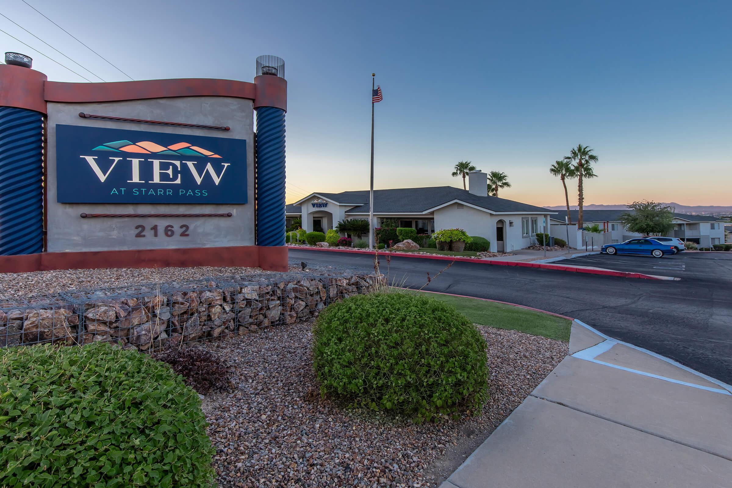 Sign for "View at Starr Pass" with the address 2162, surrounded by landscaping, palm trees, and a clear sky. The sign features a colorful mountain design and is prominently placed at the entrance to a residential area.