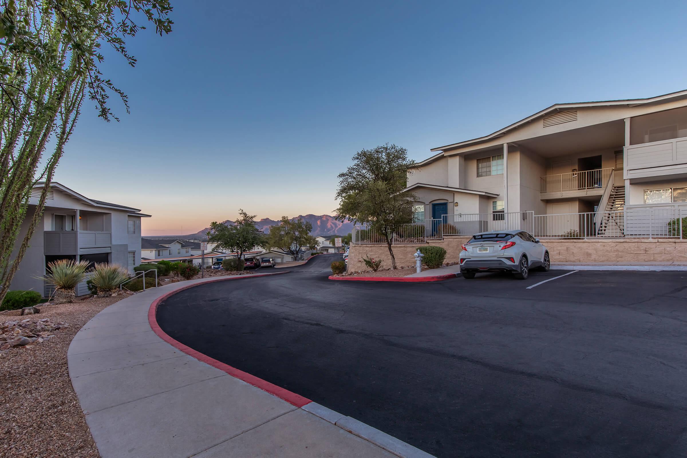 A residential area featuring a curved driveway with parked cars, surrounded by landscaped plants and trees. In the background, mountains are visible against a clear sky at dusk, creating a serene atmosphere. The architecture consists of modern two-story apartment buildings.