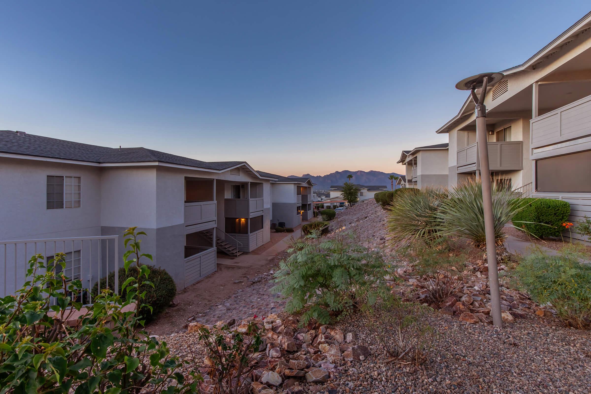 A residential area showcasing two adjacent apartment buildings with a desert landscape. In the foreground, there’s a rocky garden with desert plants, while the background features a twilight sky and distant mountains, creating a serene atmosphere.