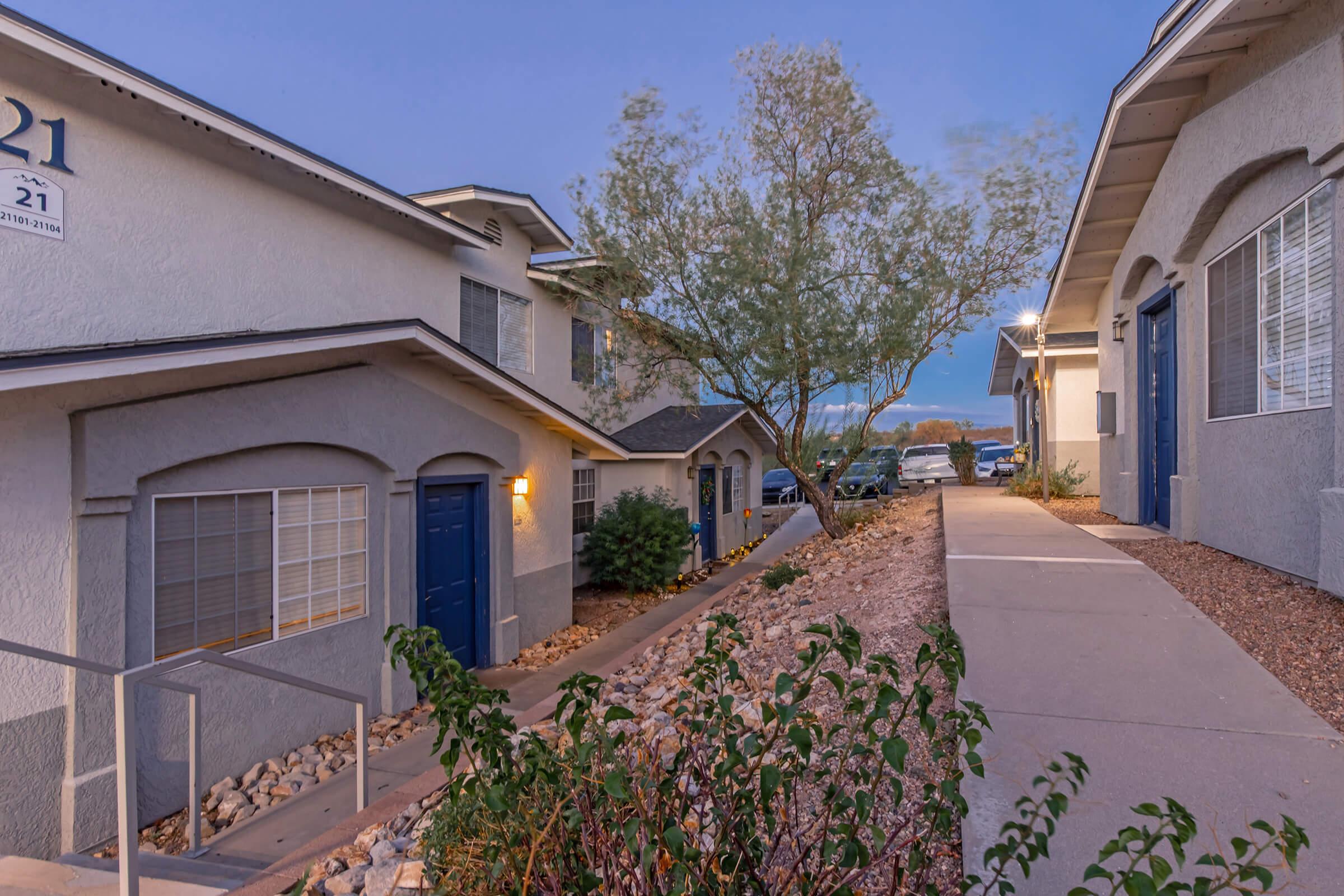 A view of two apartment buildings with blue doors, surrounded by gravel pathways and small bushes. The scene is set during twilight, with a few cars parked nearby and a tree in the background, creating a peaceful residential atmosphere.