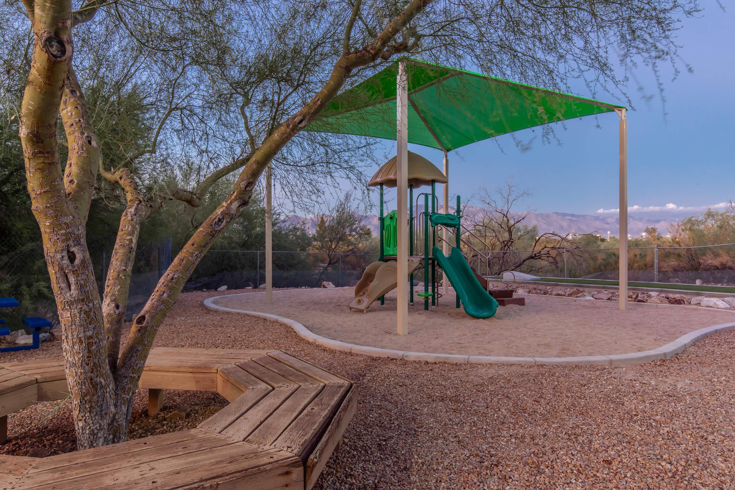 A children's playground featuring a green slide and climbing structure under a shaded canopy. The area is surrounded by gravel and has a wooden bench. In the background, mountains are visible, and the sky is a soft blue.