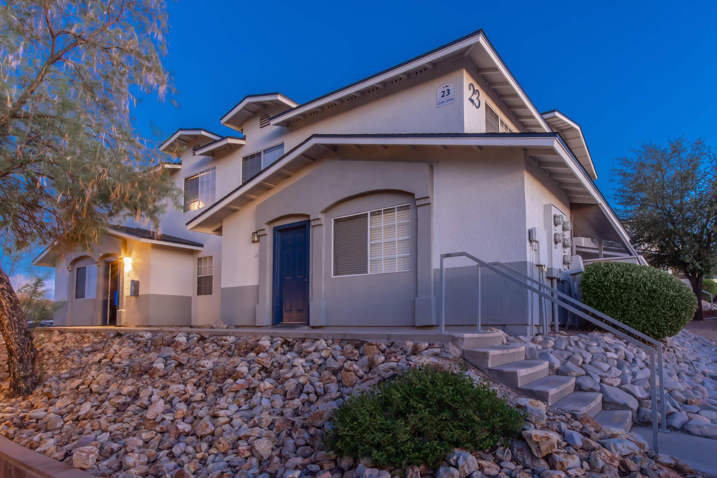 Modern two-story residential building with a grey and white exterior, set against a twilight sky. The structure features multiple windows, a blue front door, and stone landscaping with a small stairway leading to the entrance. Surrounding vegetation adds a touch of greenery to the scene.