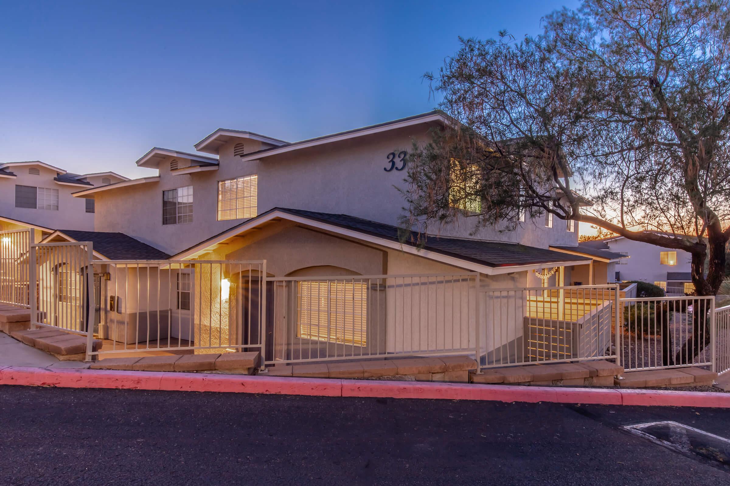 A modern residential building at dusk, featuring multiple stories with large windows. The exterior is light-colored, surrounded by a low fence and a tree nearby. The building number "33" is visible on the facade, and there are soft lights illuminating the entrance. The scene conveys a calm and inviting atmosphere.