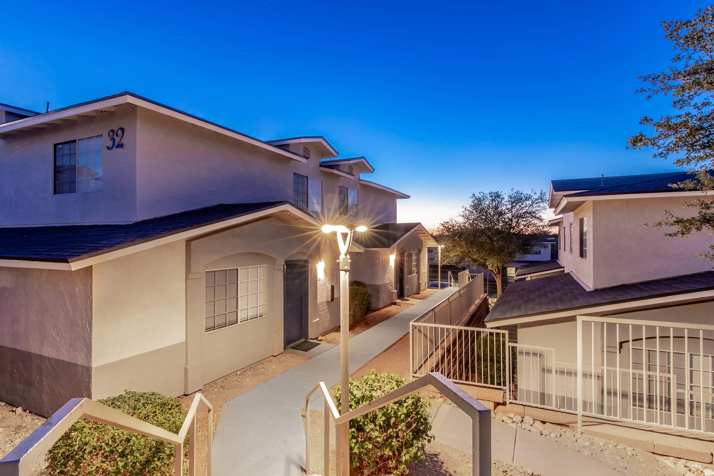 Pathway leading to residential buildings at dusk, featuring well-lit stairs and landscape shrubs. The scene captures a warm glow as the sun sets in the background, with a clear sky transitioning from blue to twilight.