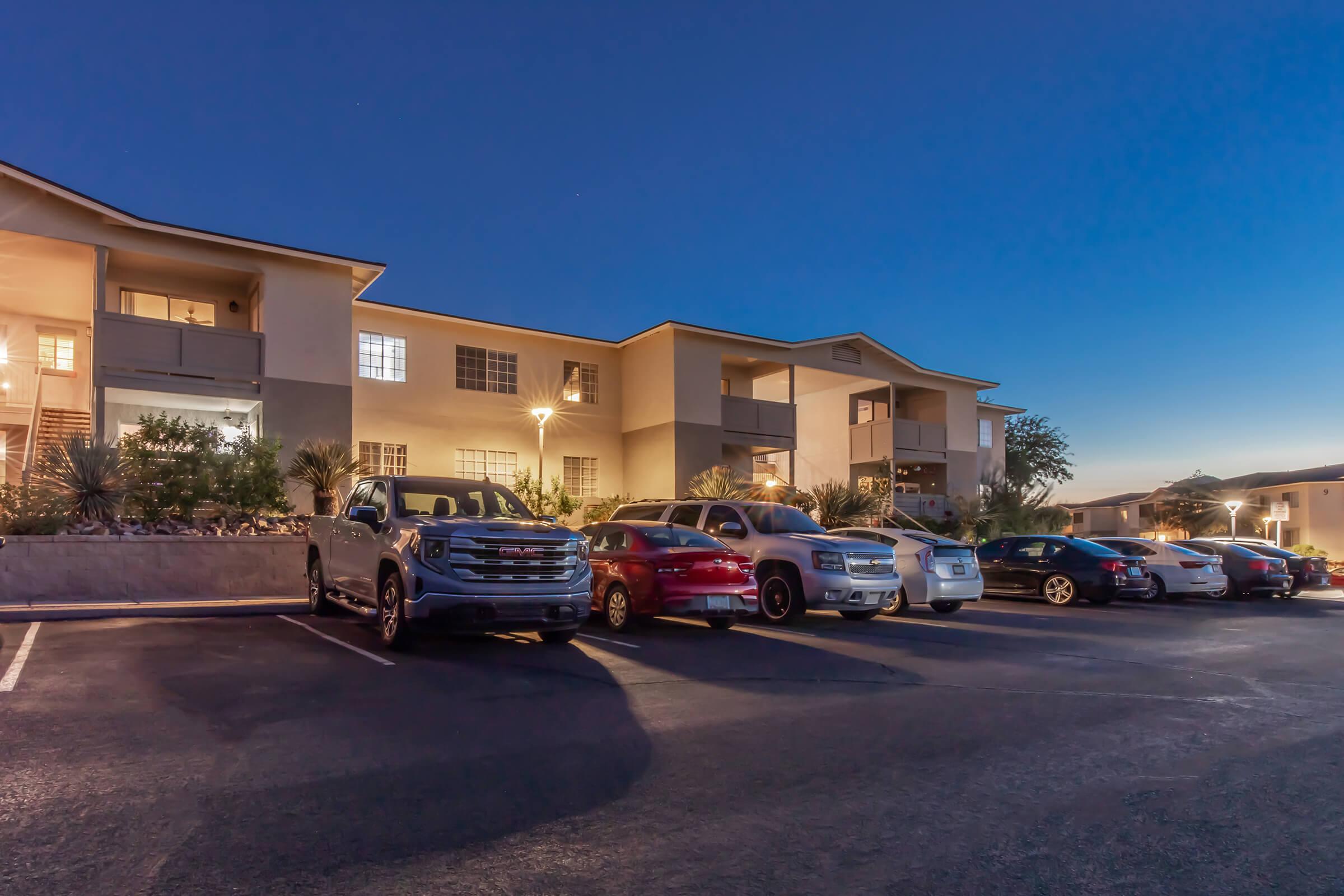 Residential apartment building at dusk, featuring multiple vehicles parked in front. The building has large windows and outdoor lighting, showing a well-maintained exterior with landscaping. The sky is transitioning from day to night, creating a serene atmosphere.
