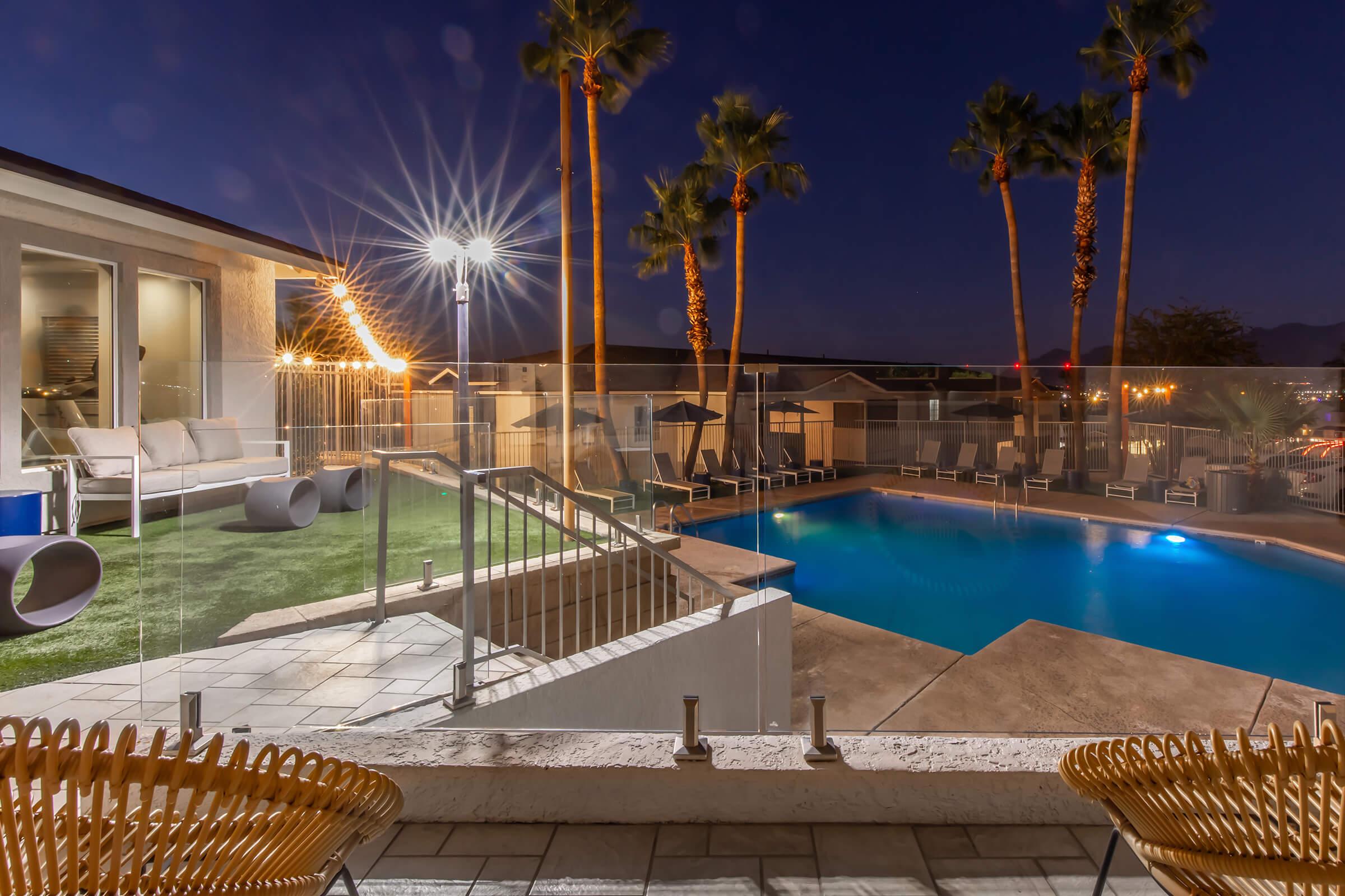 A serene nighttime view of a modern pool area, featuring a sparkling blue pool bordered by palm trees. Cozy lounge chairs are arranged poolside, with soft lighting creating a warm ambiance. The backdrop includes a stylish building with large windows.