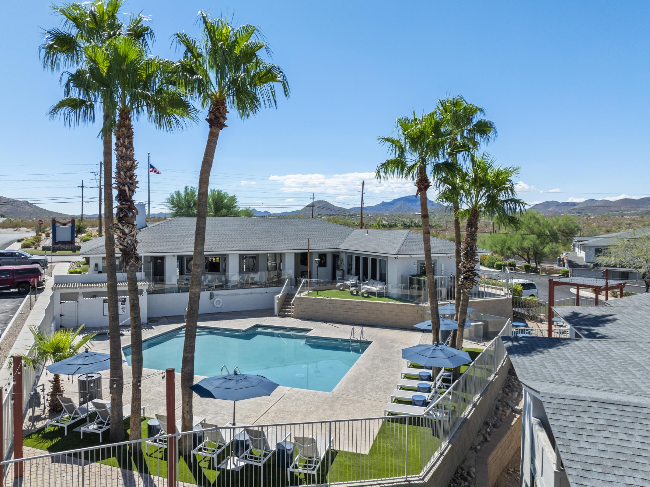 Aerial view of a resort-style pool surrounded by palm trees with lounge chairs. In the background, a modern building and mountainous landscape under a clear blue sky. The sunny setting indicates a warm climate, perfect for relaxation and leisure activities.