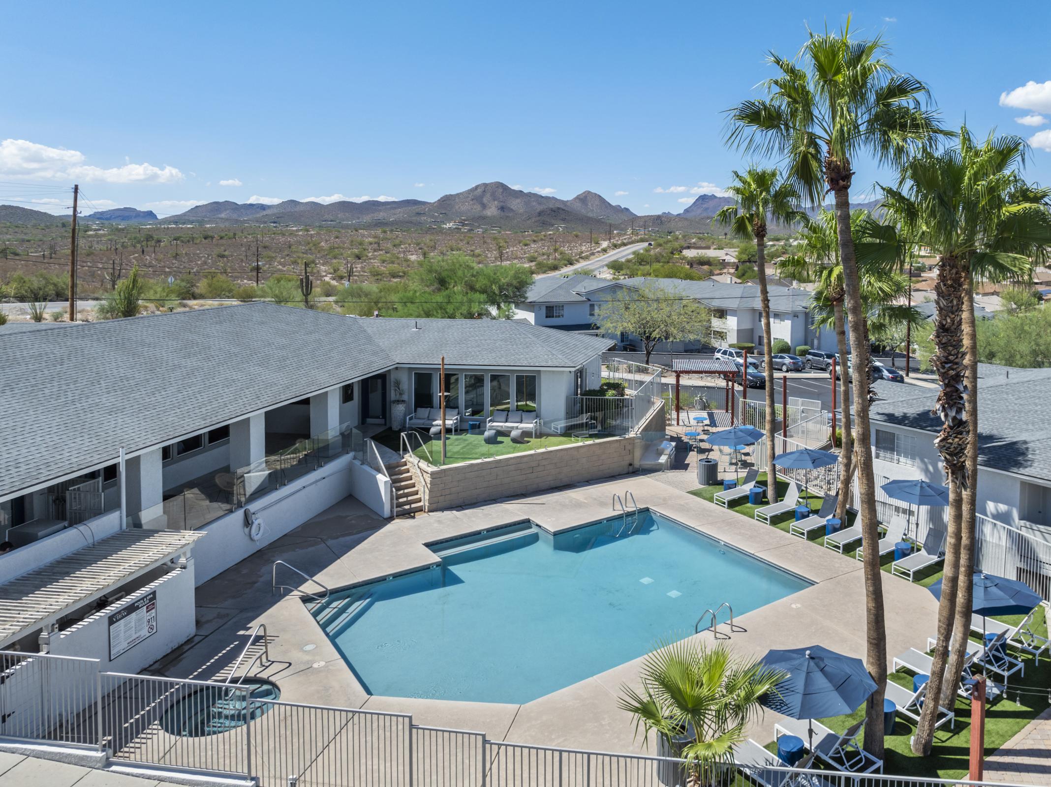 A sunny view of a hotel pool area featuring a rectangular swimming pool surrounded by lounge chairs and palm trees. In the background, there are mountains and a clear blue sky, while the hotel building is visible to the left with outdoor seating areas.
