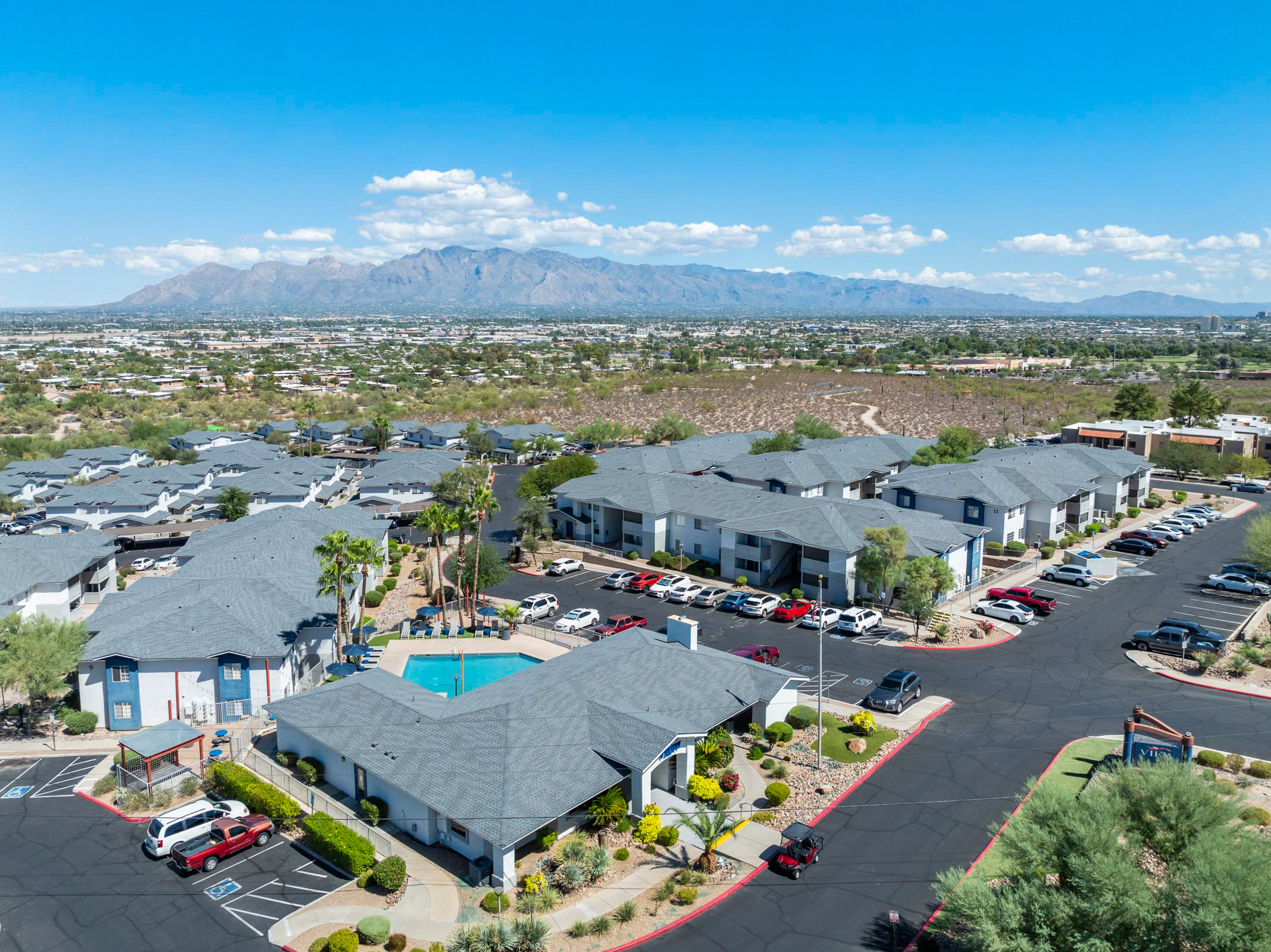Aerial view of a residential complex featuring several buildings with gray roofs, a swimming pool, and ample parking spaces. In the background, there are mountains and a clear blue sky, while the foreground includes landscaped areas with palm trees and other greenery.
