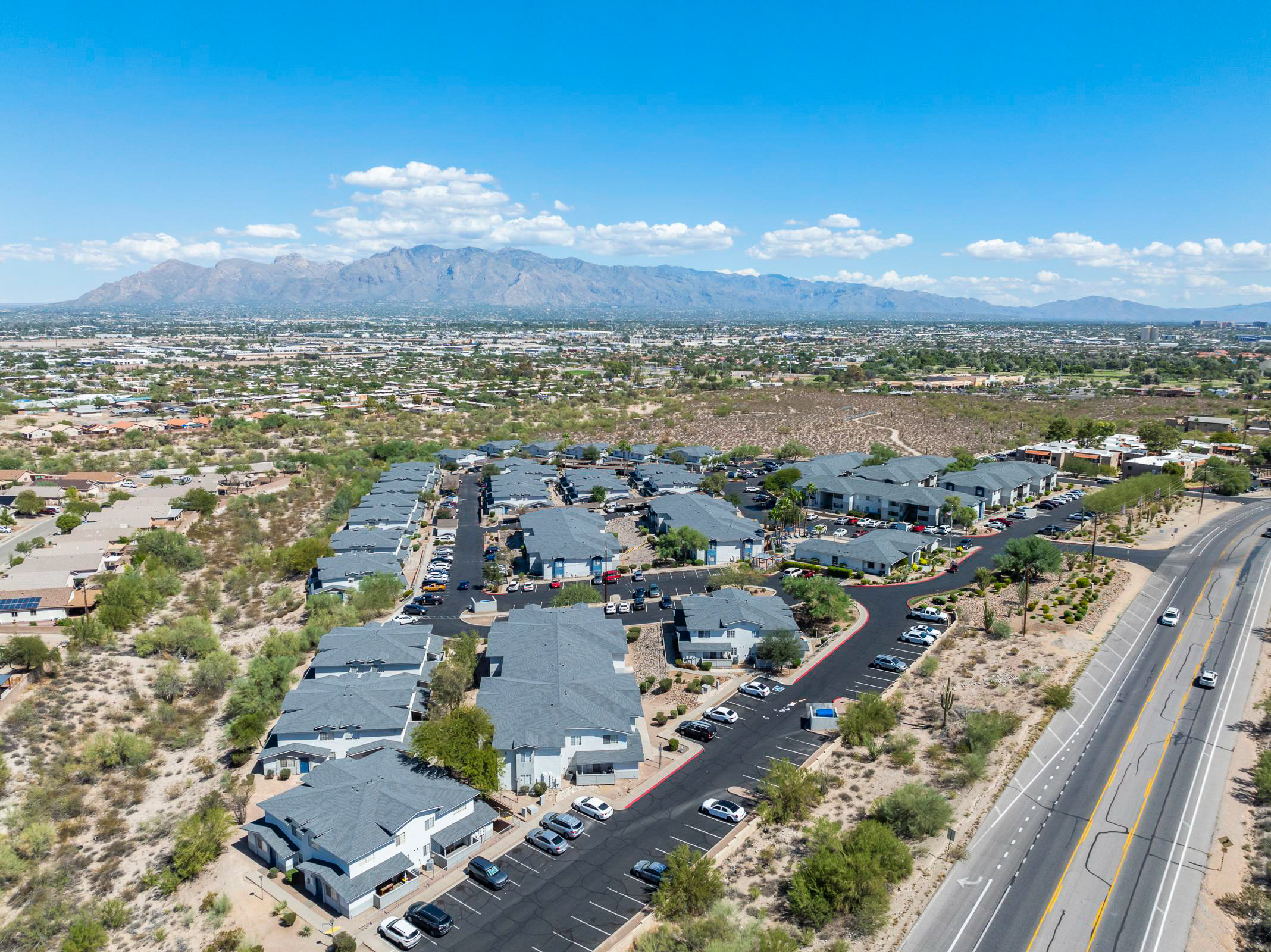 Aerial view of a suburban neighborhood featuring rows of houses with gray roofs, surrounded by desert landscape. The area includes streets filled with parked cars and a backdrop of mountains under a clear blue sky with scattered clouds.