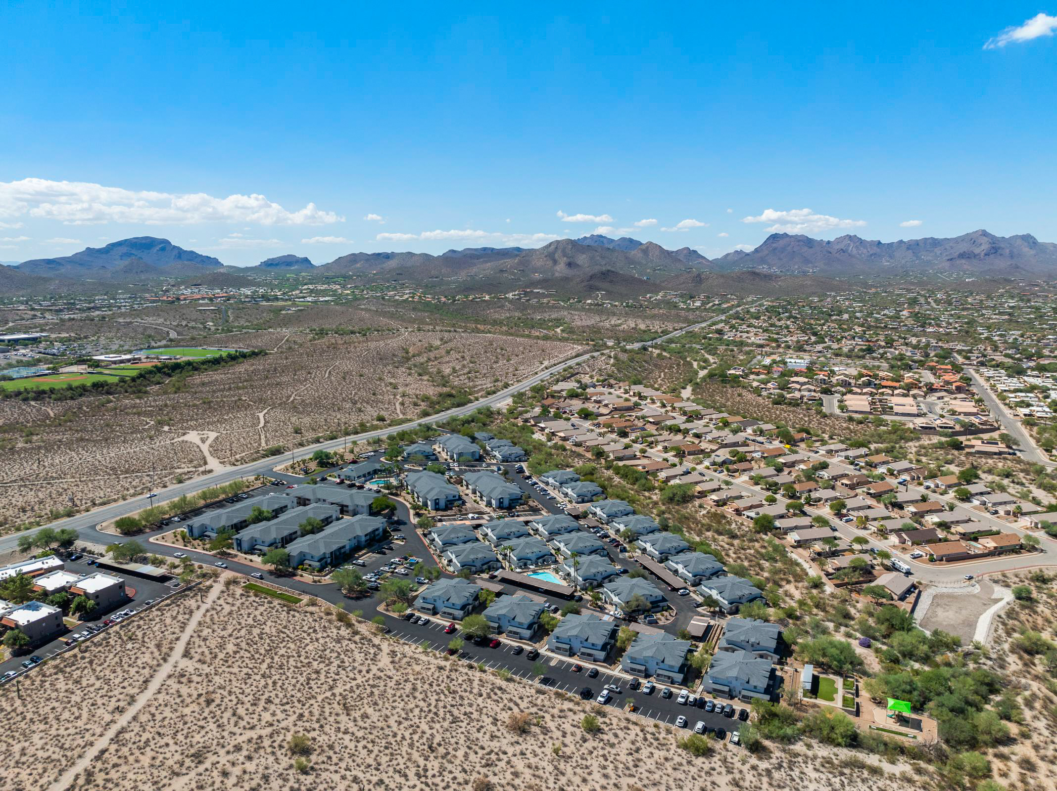 Aerial view of a residential area featuring a cluster of houses and apartment buildings surrounded by arid desert landscape and mountains in the background. Clear blue sky with a few clouds, showcasing the contrast between developed areas and natural terrain.