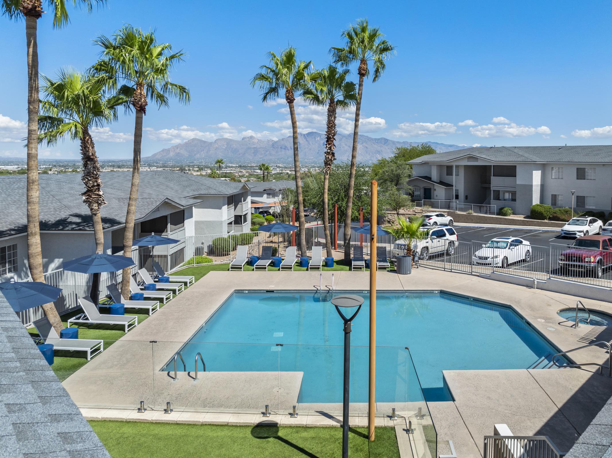 A bright outdoor swimming pool surrounded by lounge chairs and palm trees, with a view of mountains in the background. The scene features clear blue skies and modern apartment buildings nearby, creating a relaxing atmosphere.