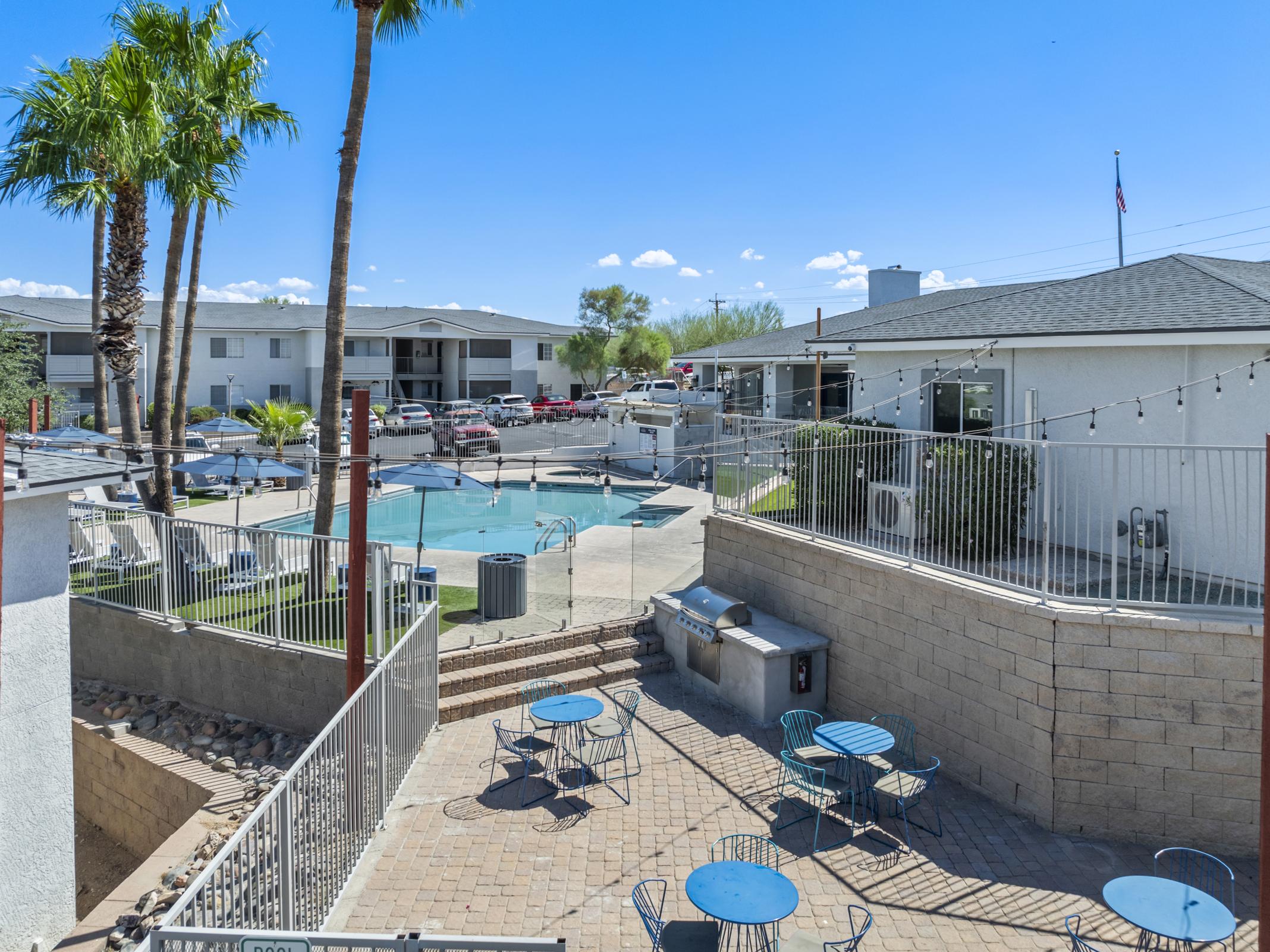 View of a residential community featuring a swimming pool surrounded by palm trees. In the foreground, there are several blue tables and chairs on a patio area. The background shows parked cars and buildings, with a clear blue sky above. The scene conveys a sunny, inviting atmosphere.