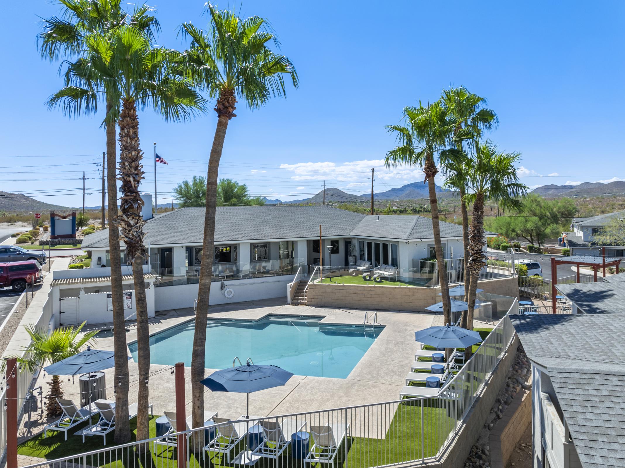 A sunny view of a hotel pool area surrounded by palm trees. Lounge chairs are positioned around the pool, and a building with large windows overlooks the scene. Blue skies and mountains in the background enhance the resort-like atmosphere.