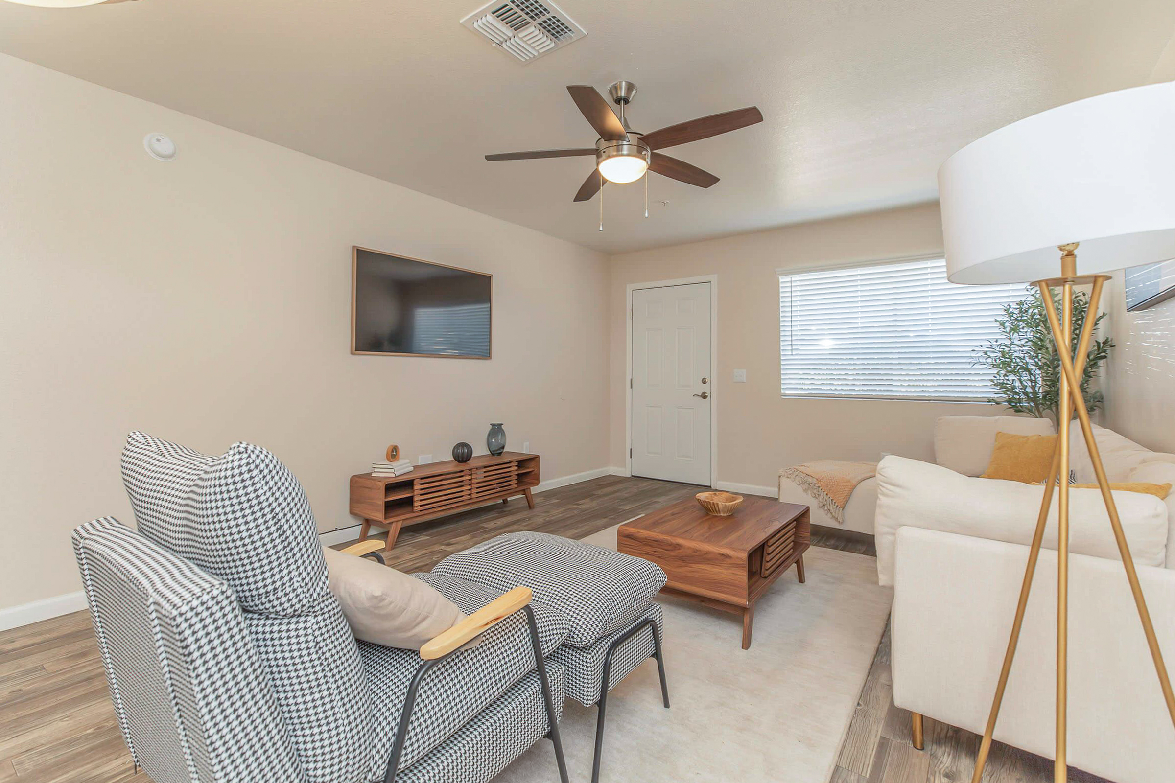 A modern living room featuring a light color scheme. There are two comfortable chairs, a wooden coffee table, and a cozy sofa decorated with pillows. A flat-screen TV is mounted on the wall, and a door and window allow natural light to fill the space. The flooring is laminate, enhancing the room's contemporary look.