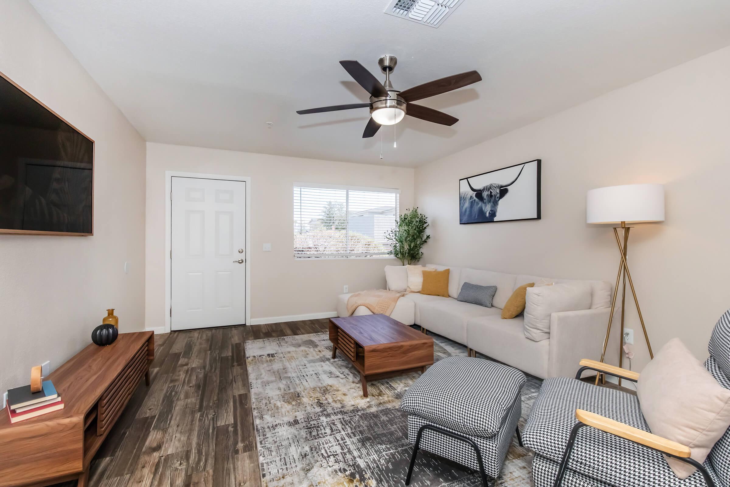 A modern living room featuring a light-colored couch with colorful throw pillows, a wooden coffee table, and stylish armchairs. There's a flat-screen TV mounted on the wall, a large window providing natural light, and a door leading outside. Decorative elements include a lamp and artwork on the wall.