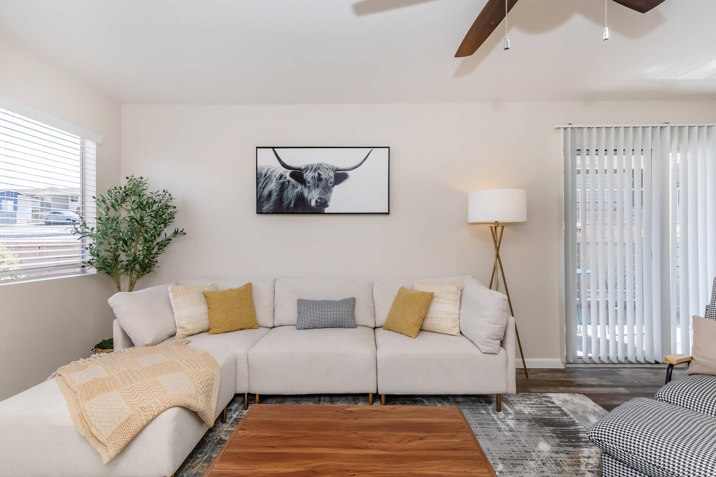 A cozy living room featuring a large, light-colored sectional sofa with decorative pillows, a wooden coffee table, and a potted plant. Above the sofa, there is a framed artwork of a bull. The room has natural light from a window, and there are vertical blinds and a floor lamp in the corner.
