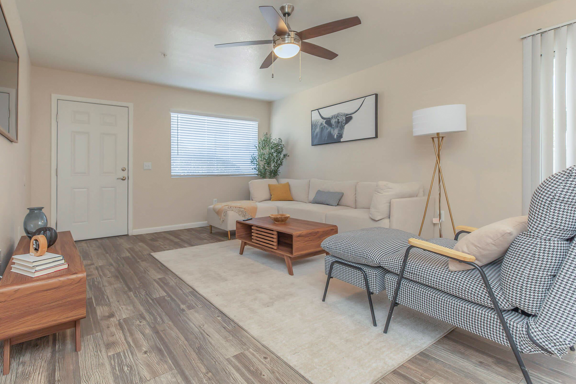 Modern living room featuring a light-colored couch, wooden coffee table, and patterned armchair. A ceiling fan hangs above, and a potted plant adds greenery. The walls are neutral-toned, and there’s a window with blinds. Decor includes a framed artwork and a lamp, creating a cozy and inviting atmosphere.
