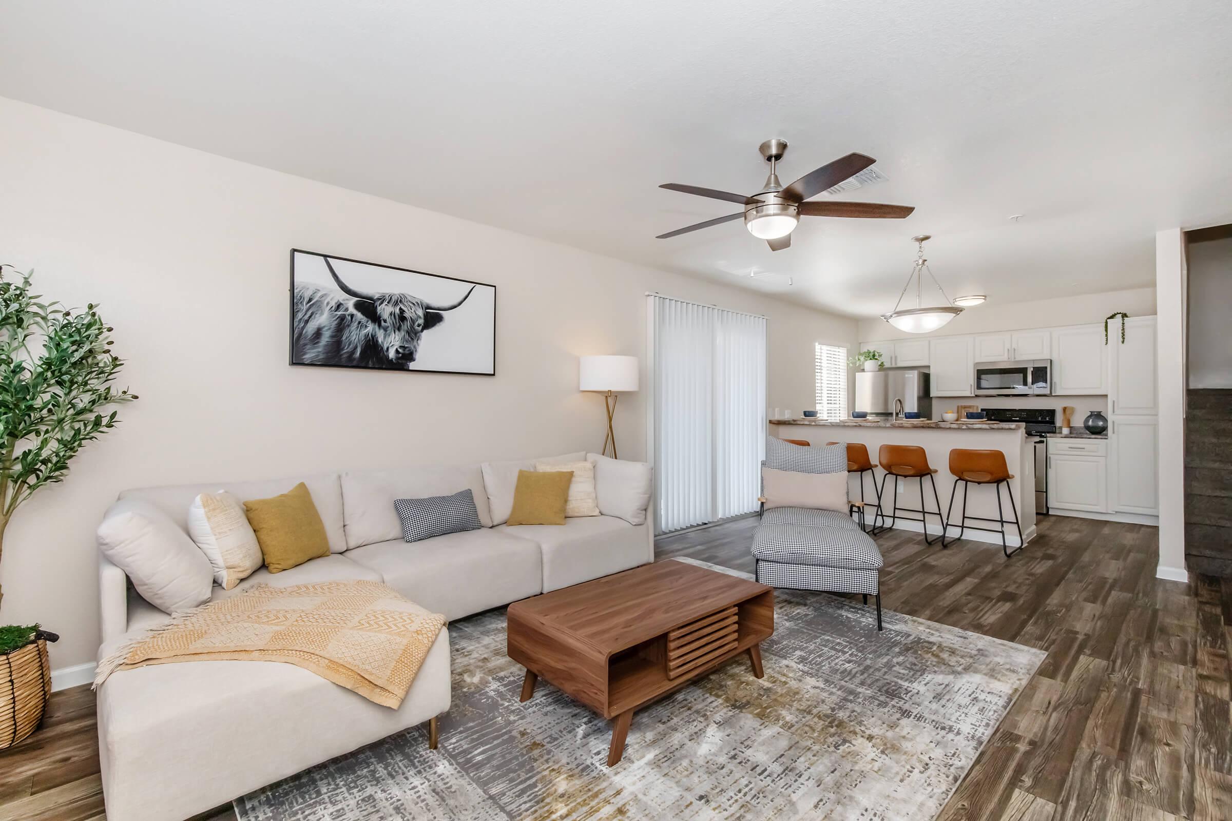 Modern living room with a light-colored sectional sofa and decorative pillows. A wooden coffee table sits on a patterned rug, and there’s a large cow portrait on the wall. To the side, a kitchen area with bar stools is visible, complemented by a ceiling fan and natural light from a window.