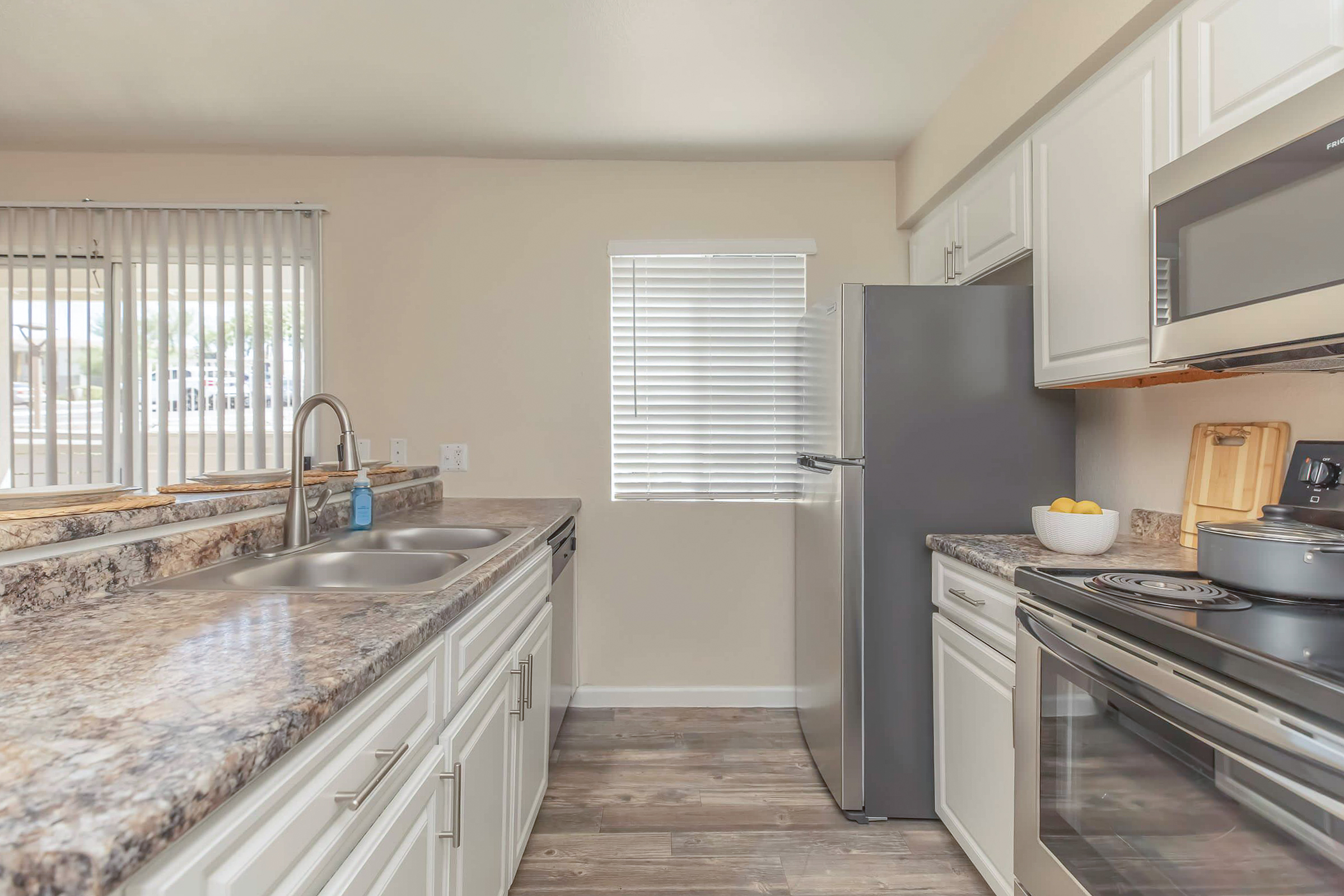 Light and modern kitchen featuring a double sink, stainless steel refrigerator, and an oven with a microwave above. The countertops are a mix of gray and white granite, with white cabinetry. Window blinds allow natural light, creating a bright and inviting atmosphere.