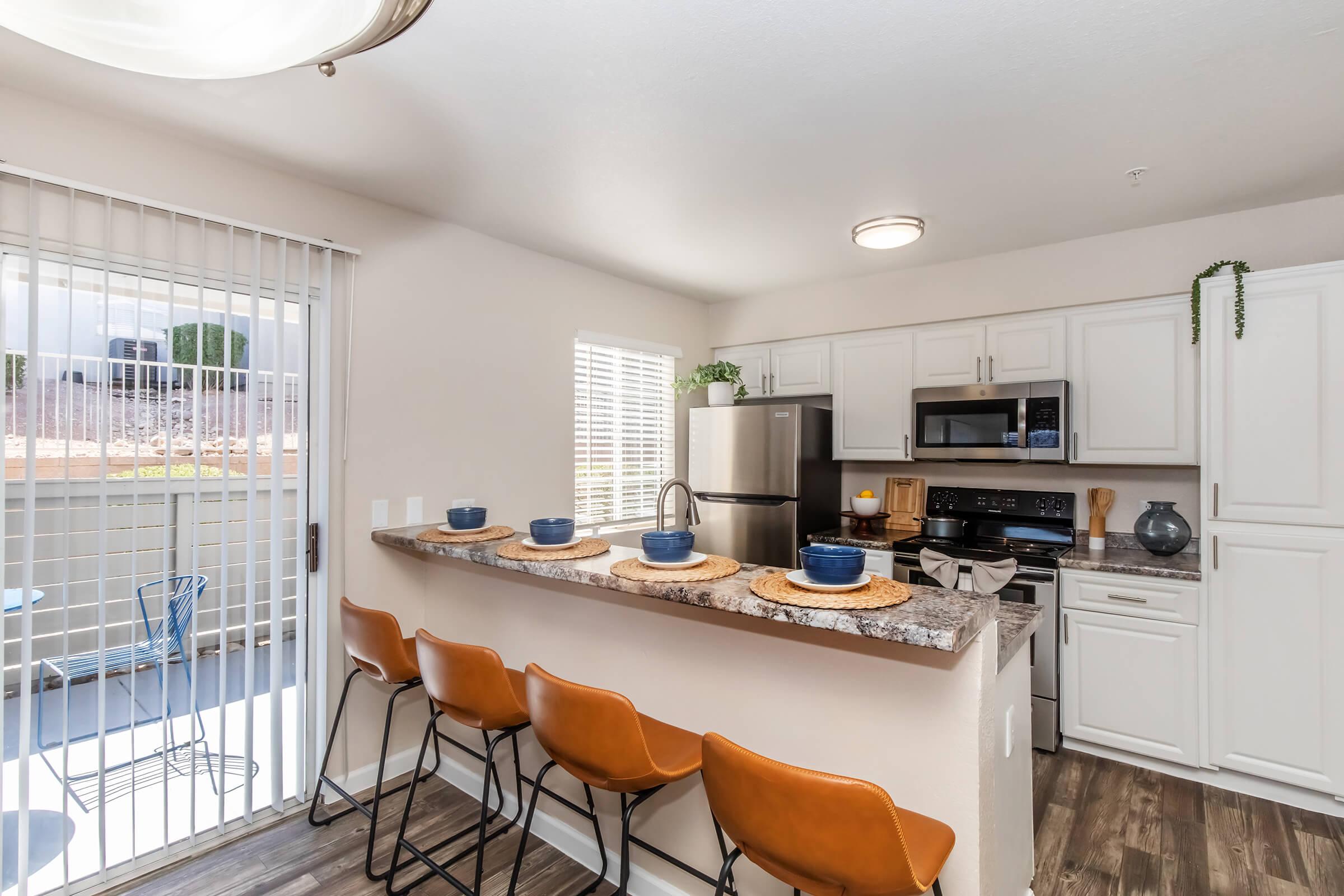 Modern kitchen with a granite countertop and bar seating for four. Features stainless steel appliances, including a fridge and microwave. Light-colored cabinets, a large window with blinds, and a sliding glass door lead to an outdoor space. Blue dishes are set on the counter, enhancing the inviting atmosphere.