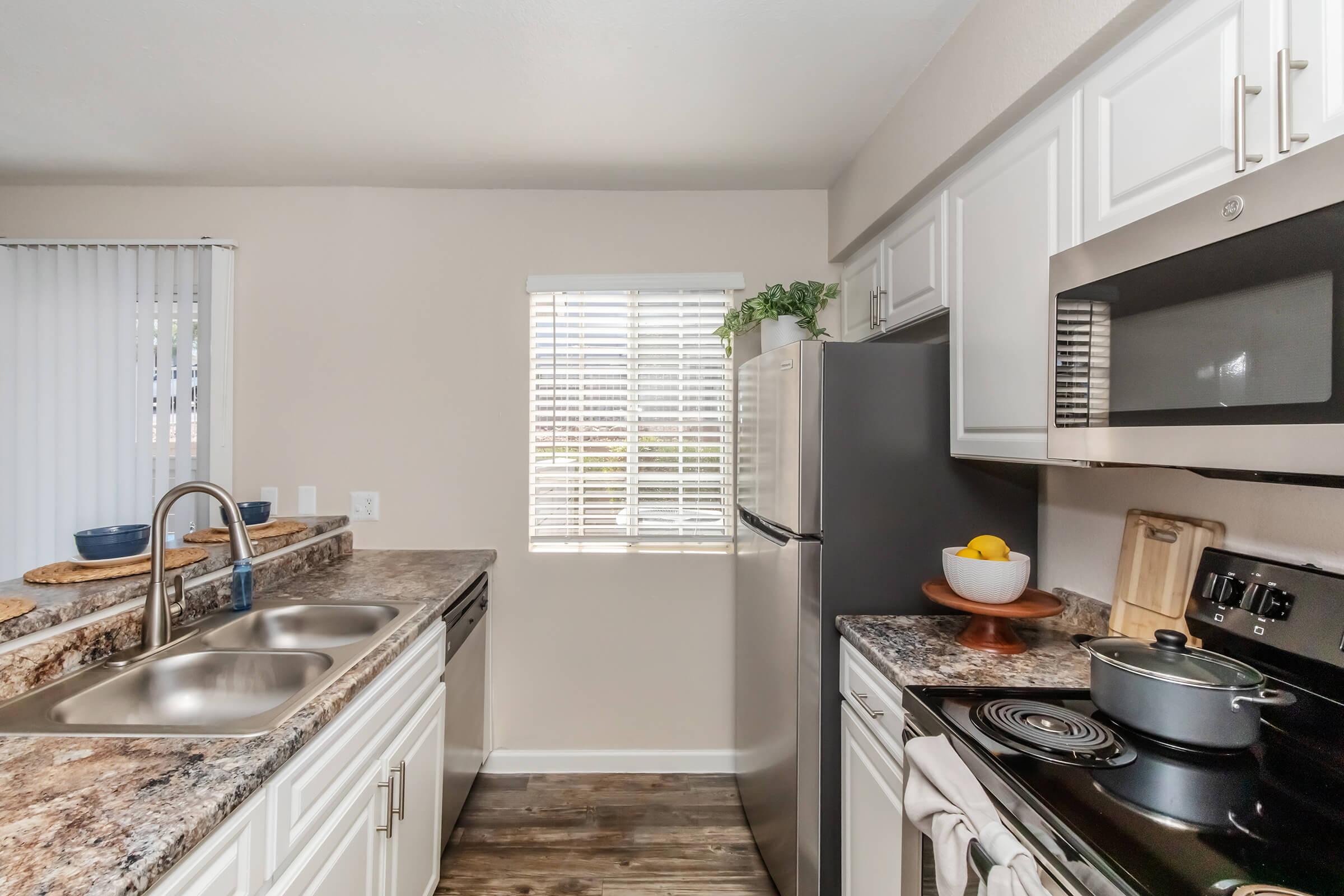 A modern kitchen featuring stainless steel appliances, including a refrigerator and stove. The countertops are made of granite, with a double sink and wooden cutting board. A window is partially opened, allowing natural light to illuminate the space, complemented by decorative items on the counters.