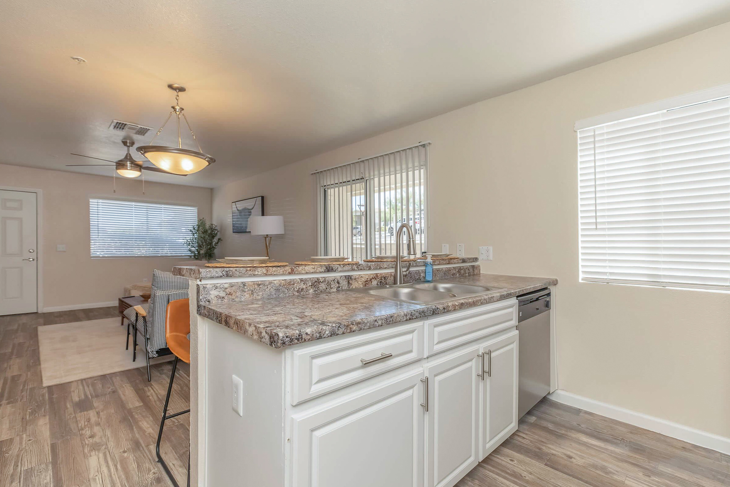 Modern kitchen with a large granite countertop and stainless steel sink. In the background, there's a cozy living area with a sofa, chairs, and a lamp. Natural light comes in through window blinds, and the space features neutral-colored walls and wood-like flooring.