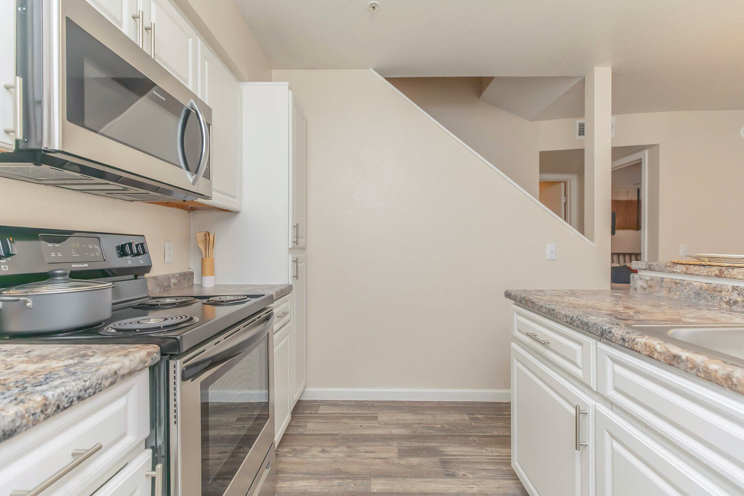 A modern kitchen featuring stainless steel appliances, including a microwave and oven. The countertops are granite, and there are white cabinets. A staircase is visible in the background, leading to an upper level. The floor is a wood-like laminate, creating a warm, inviting atmosphere.