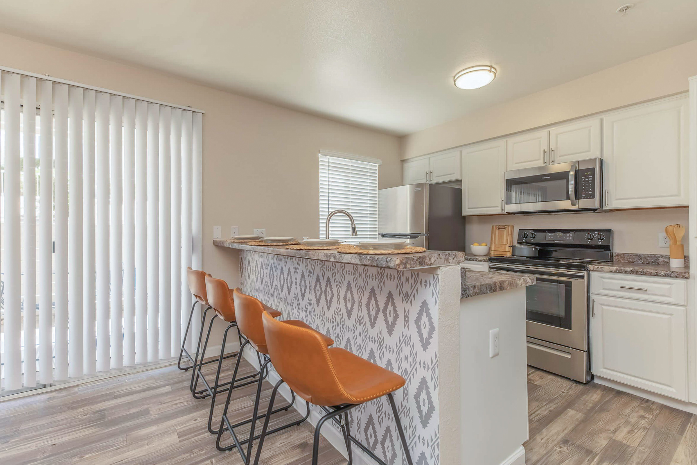 Modern kitchen featuring a countertop bar with three brown leather stools, stainless steel appliances including a refrigerator and microwave, white cabinetry, and a sliding glass door with blinds allowing natural light. The decor includes a patterned backsplash and wood-style flooring.