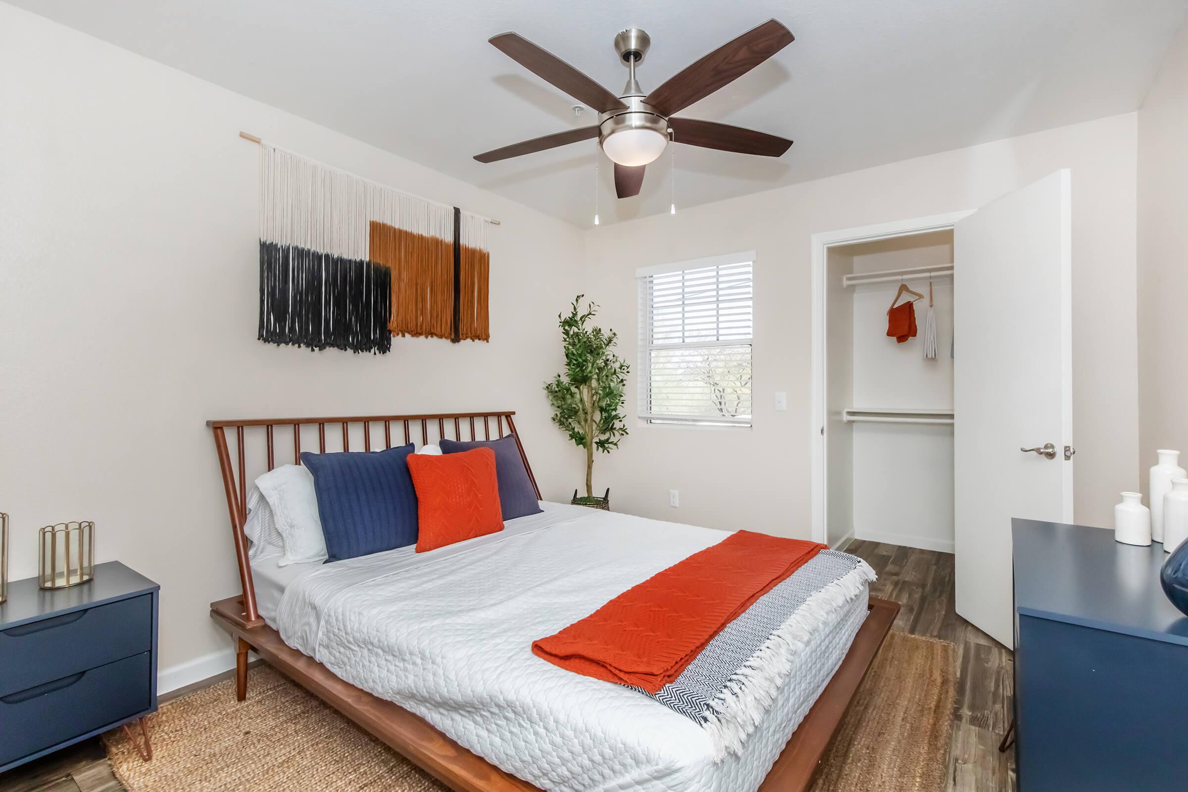 A cozy bedroom featuring a wooden bed with white bedding and colorful accent pillows. There is a decorative wall hanging, a ceiling fan, and a small plant. A blue nightstand and a closet with an open door are visible, enhancing the modern and inviting atmosphere of the space.